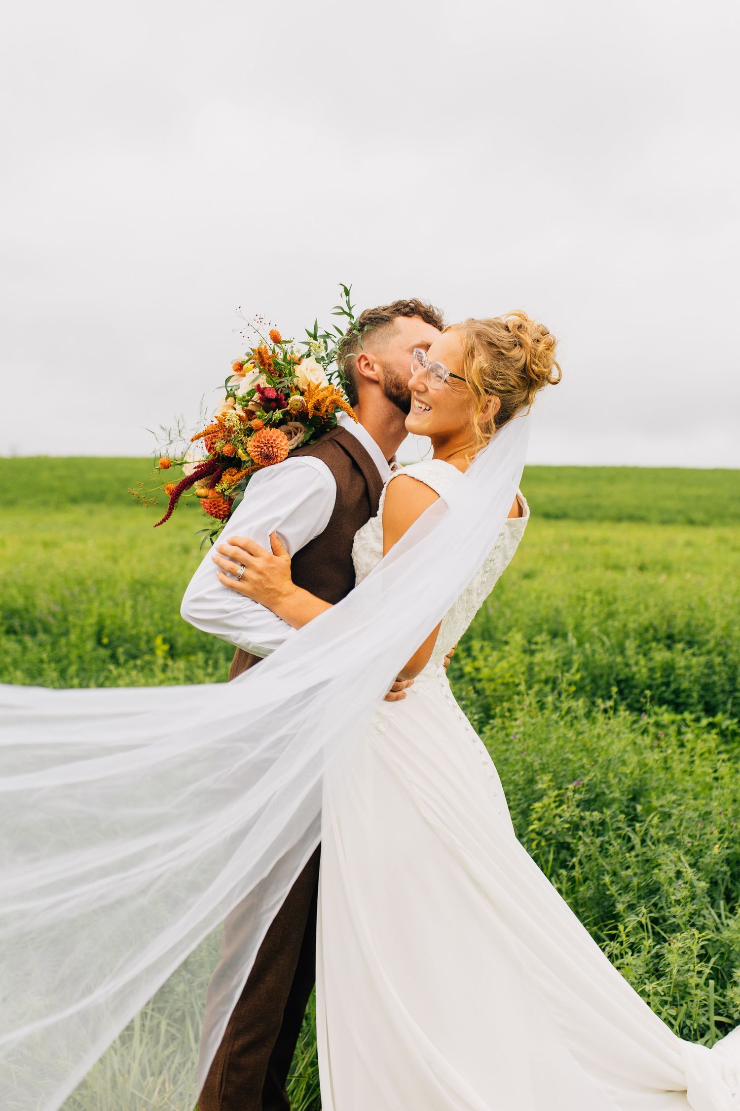 Wedding couple embraces in field as bride's veil flows dramatically in the wind.