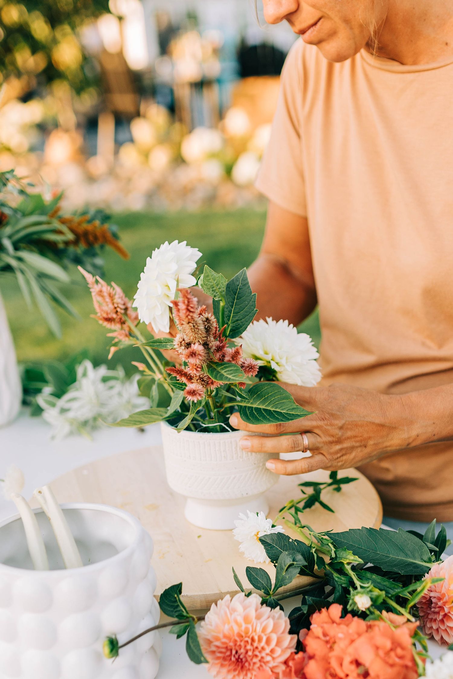 Close-up of hands arranging white and peach flowers in a decorative vase outdoors.