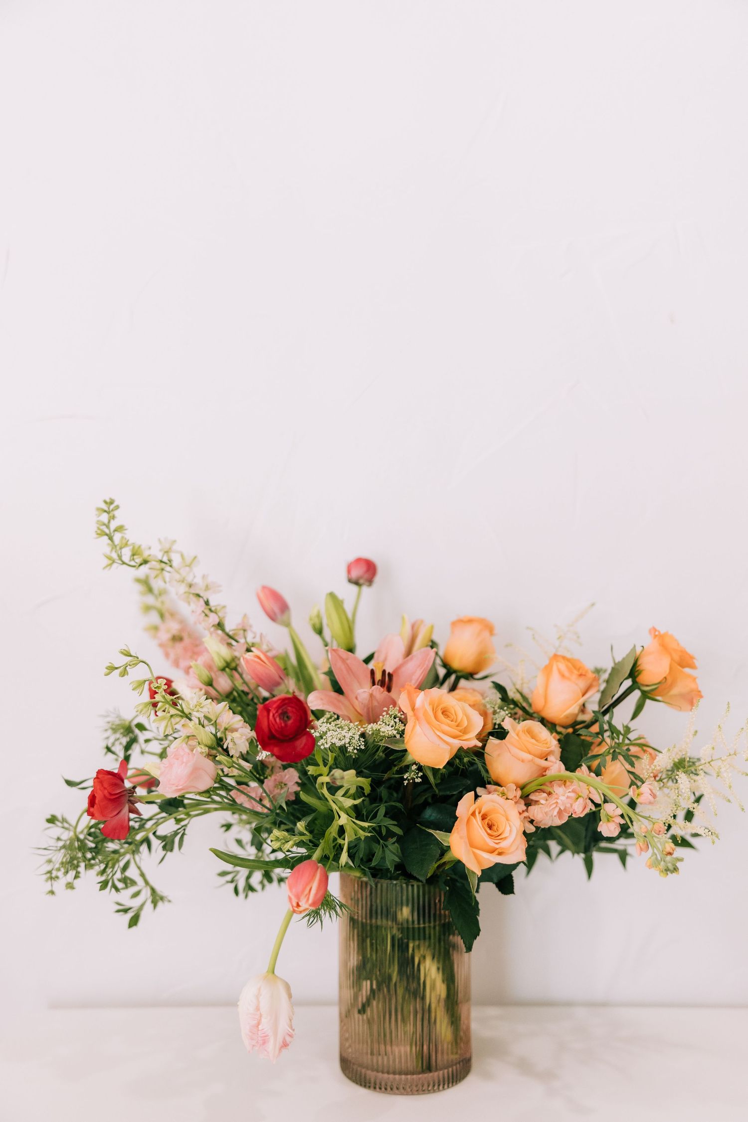 A romantic floral arrangement with peach roses, red blooms and greenery in a ribbed glass vase against a light background.