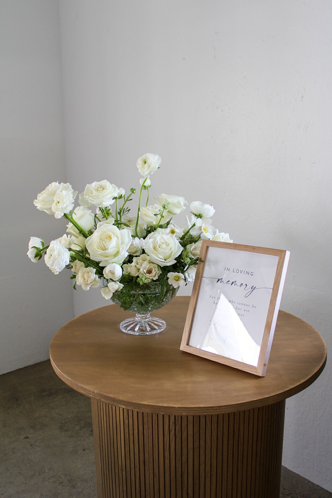 White roses in a crystal vase sit atop a round wooden pedestal table with ribbed base and framed cards.