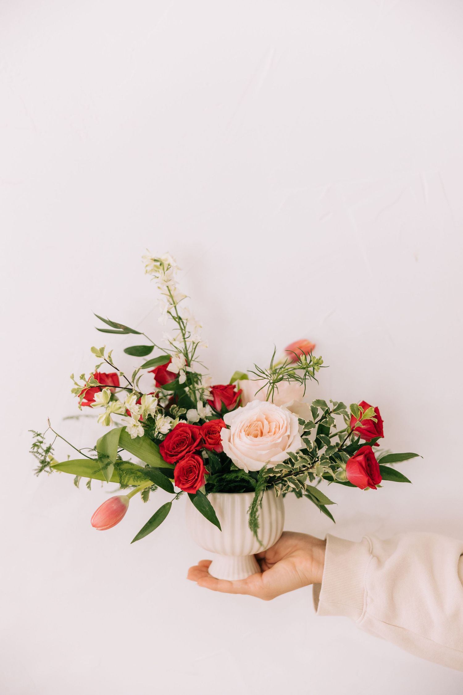 Delicate floral arrangement with red roses and peach blooms in white ceramic vase held against pink wall.