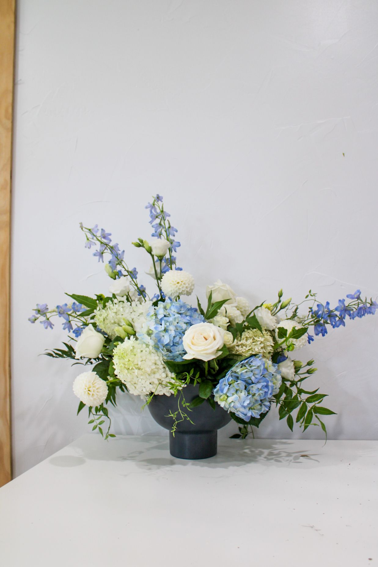 Fresh arrangement of white roses, hydrangeas and blue delphiniums in dark container on white surface.