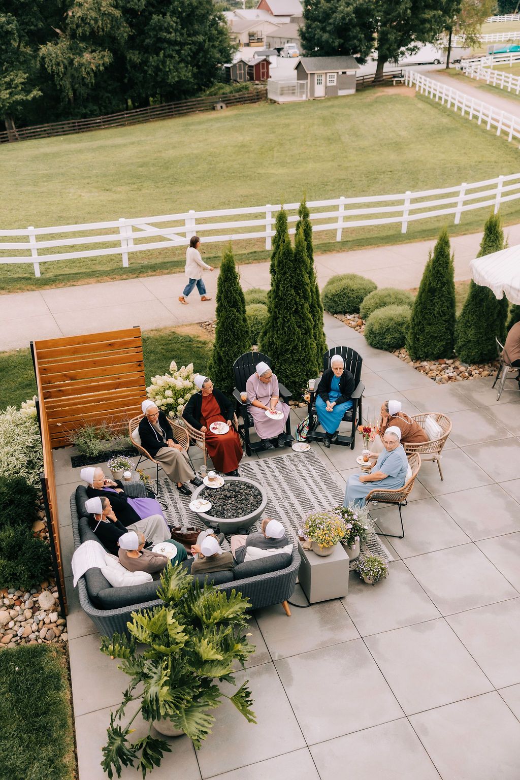 People gather on outdoor benches in a garden patio area with white fencing and landscaping in the background.