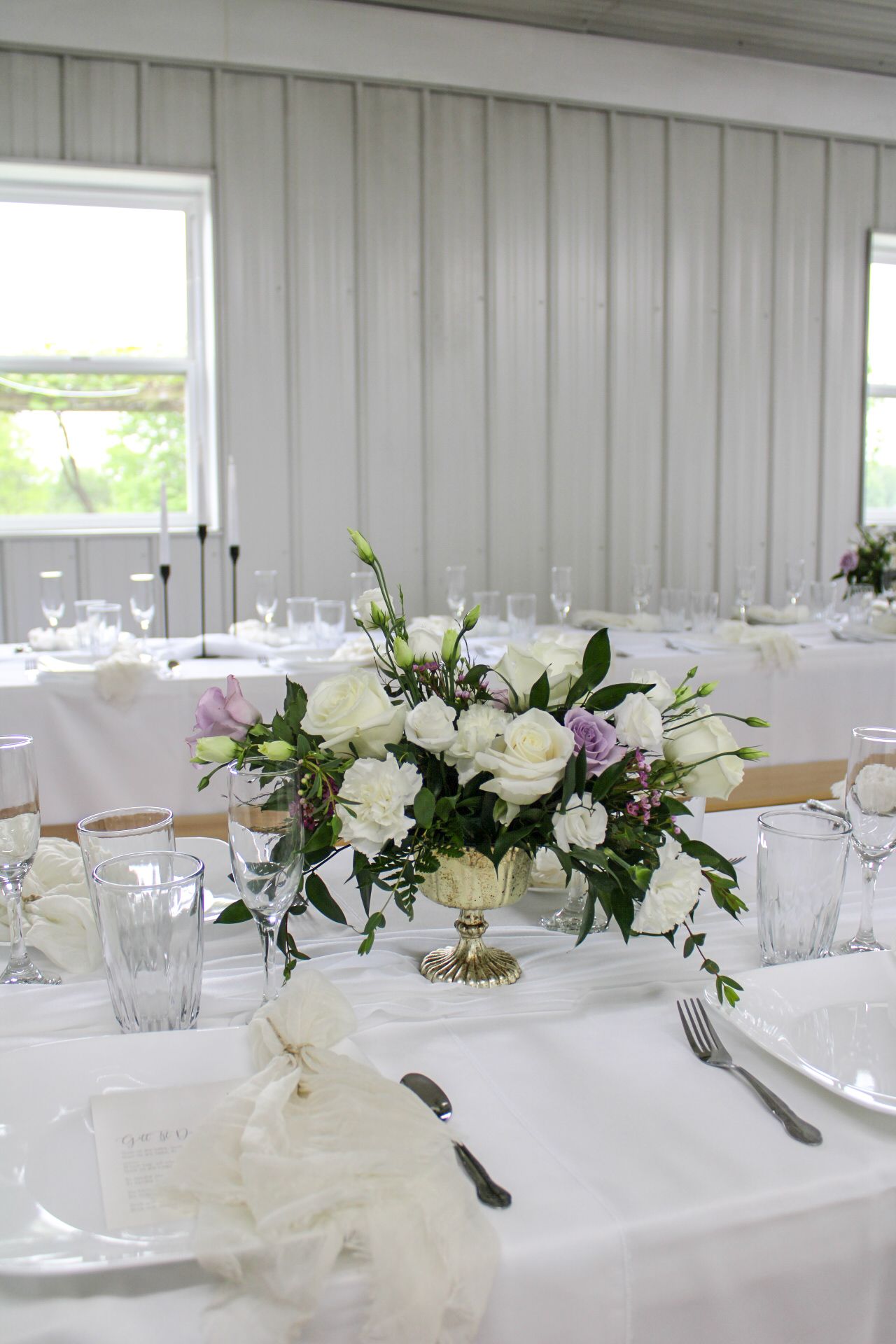 White roses and greenery centerpiece in a gold vase on crisp white tablecloth with formal place settings.