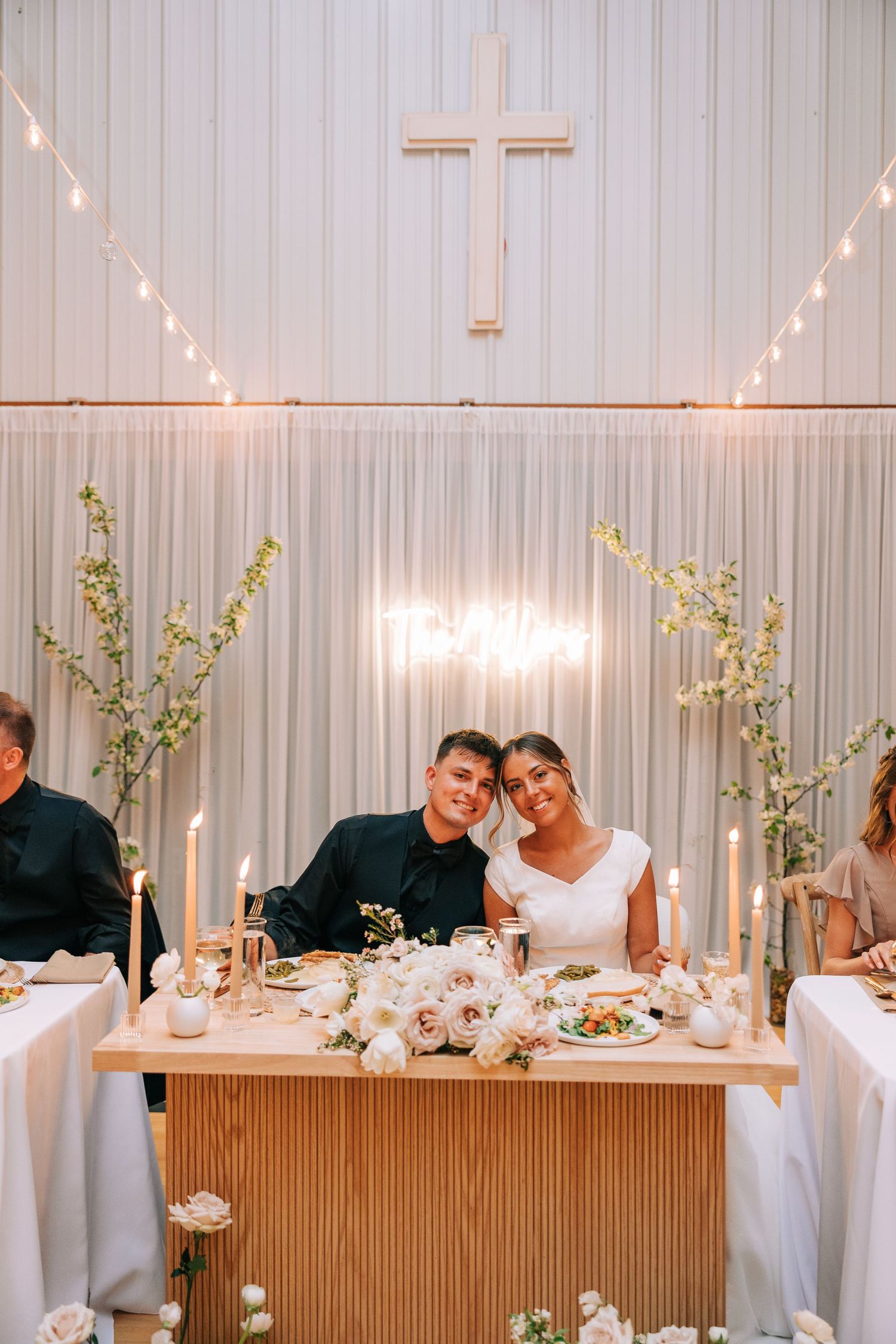 Wedding reception table under cross and string lights with elegant white drapery and floral decor.