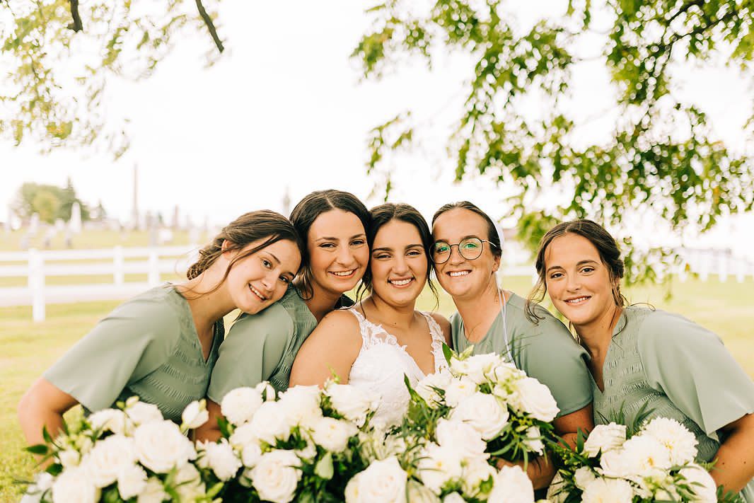 Bridesmaids in sage green dresses gather close with white floral arrangements under tree branches.