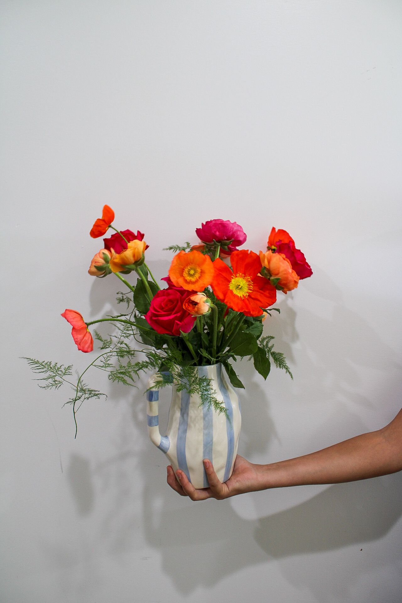 Hand holding a striped ceramic pitcher vase with vibrant red, orange, and pink poppies and ranunculus bouquet against white wall.