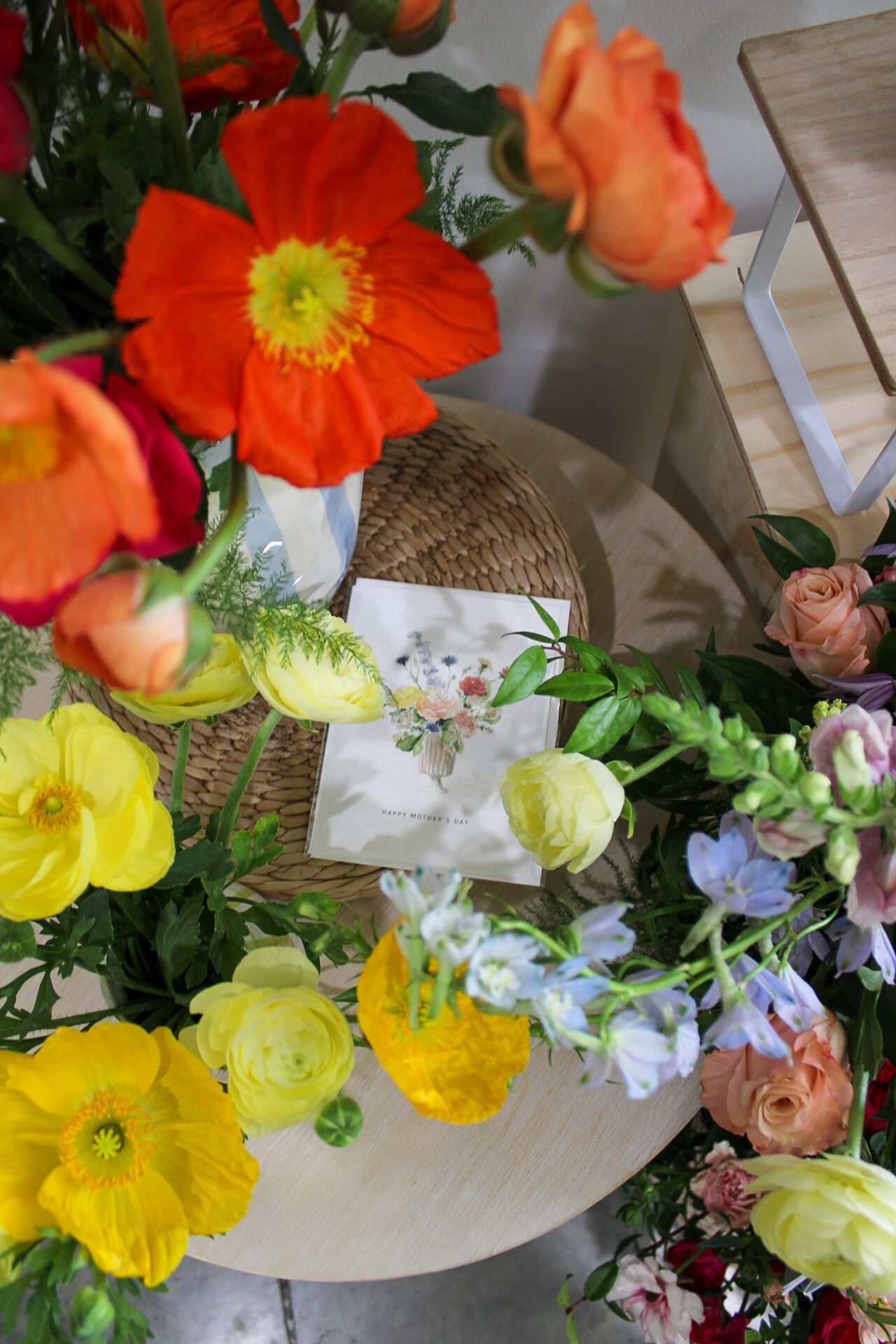 Vibrant floral arrangement featuring orange poppies, yellow ranunculus, and pink roses on a wooden table with a greeting card.