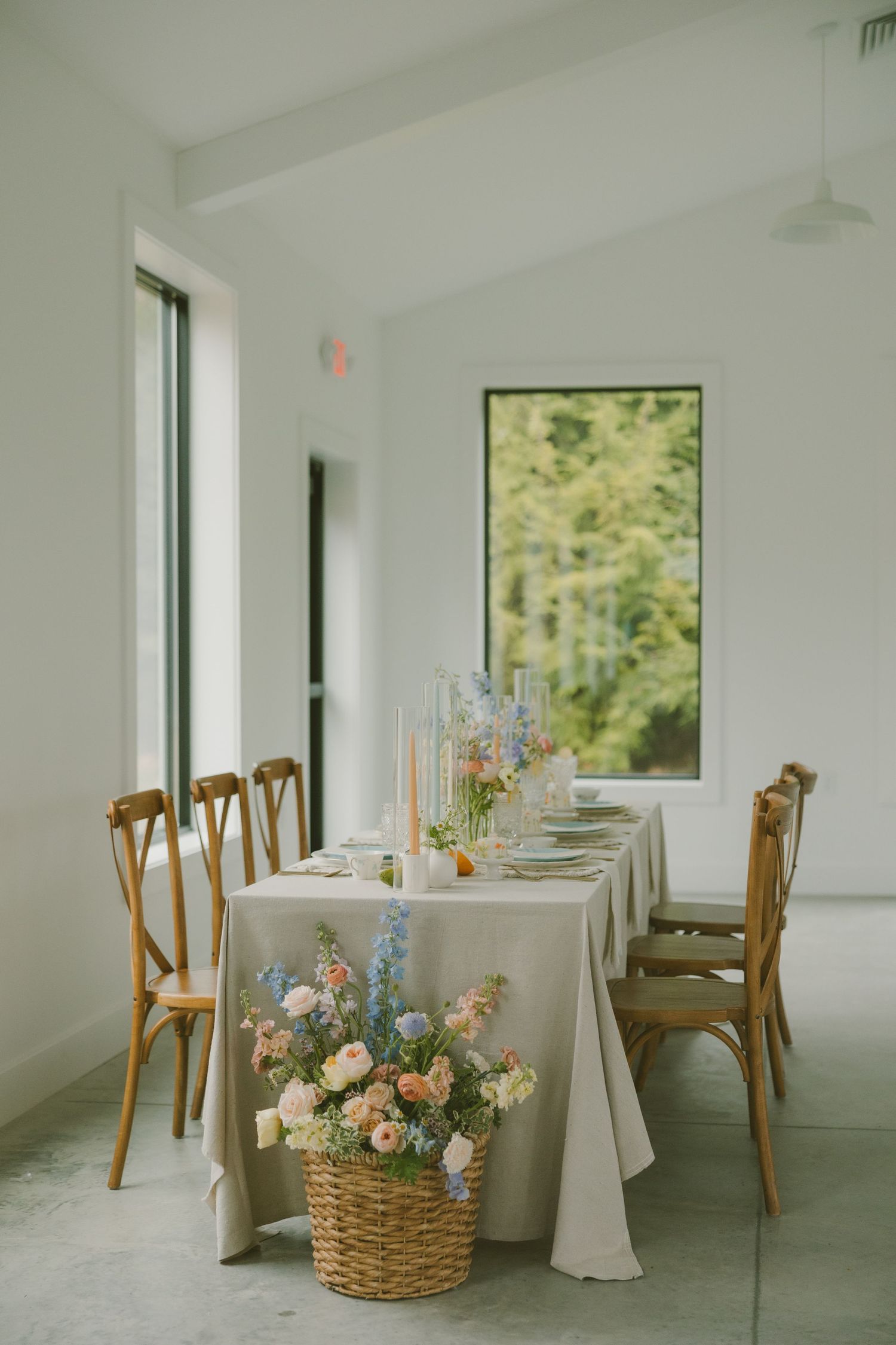 Elegant wedding reception table with wooden chairs and floral arrangements in a bright white room with windows.