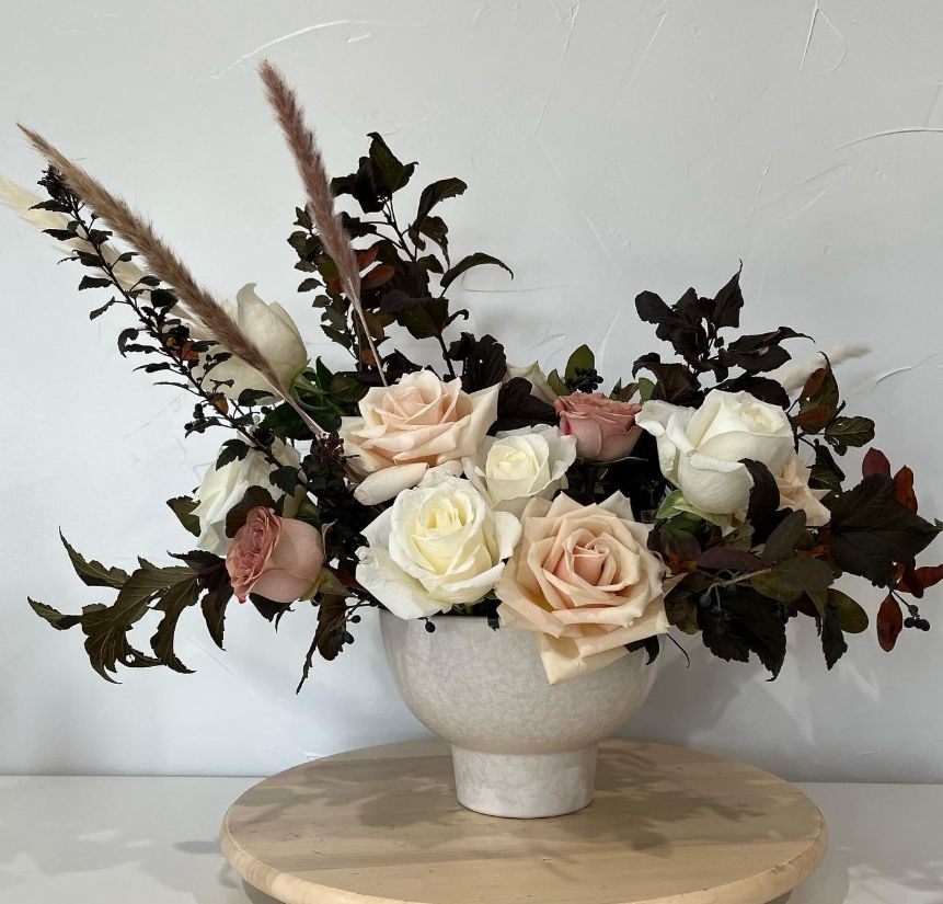 Romantic arrangement of cream and blush roses with dark foliage in a textured white bowl.