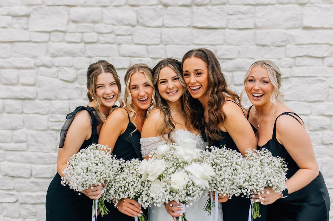 Bridesmaids in navy dresses holding baby's breath bouquets pose against white brick wall.