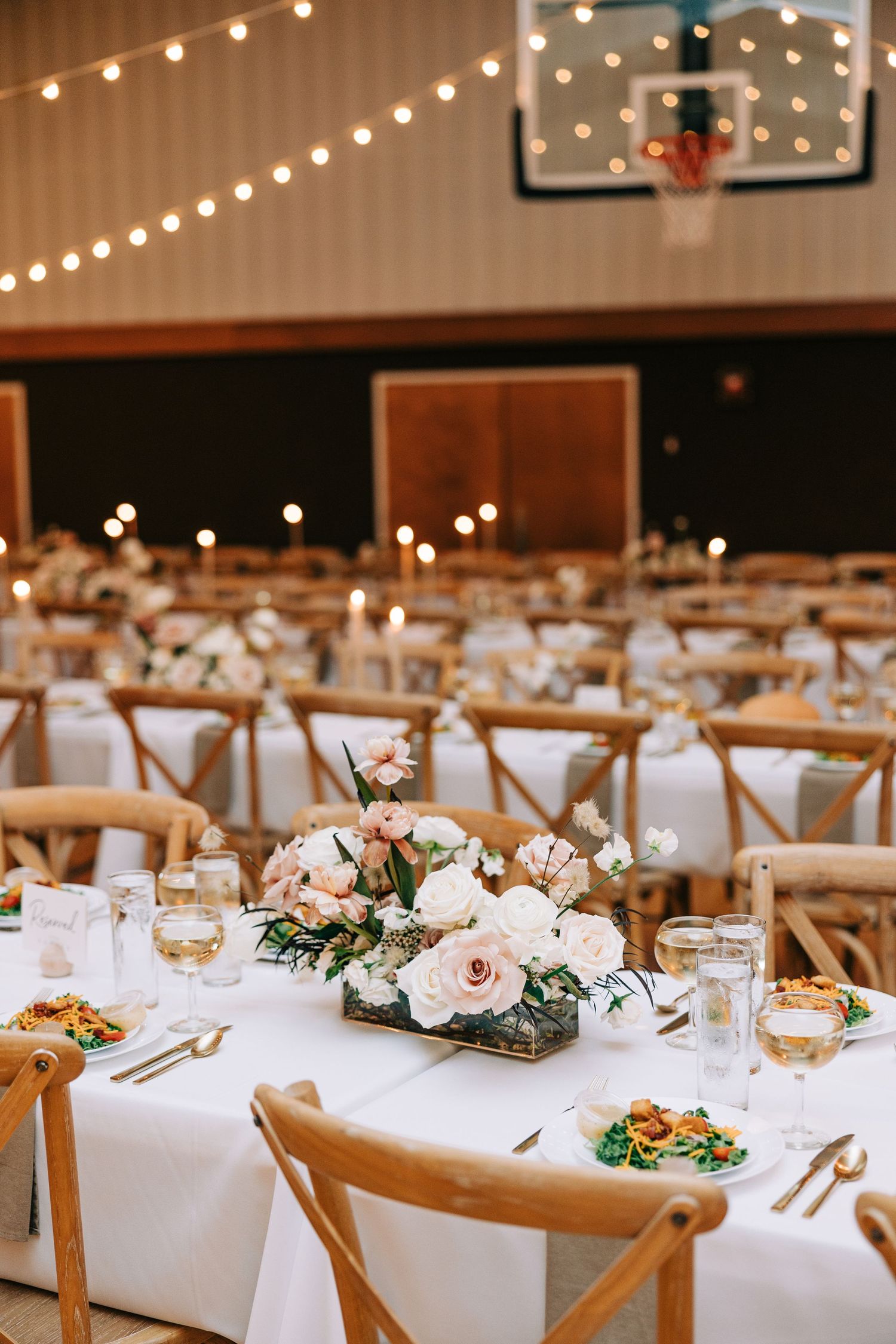 Wedding reception in a gymnasium with string lights and cross-back chairs around white-clothed tables.