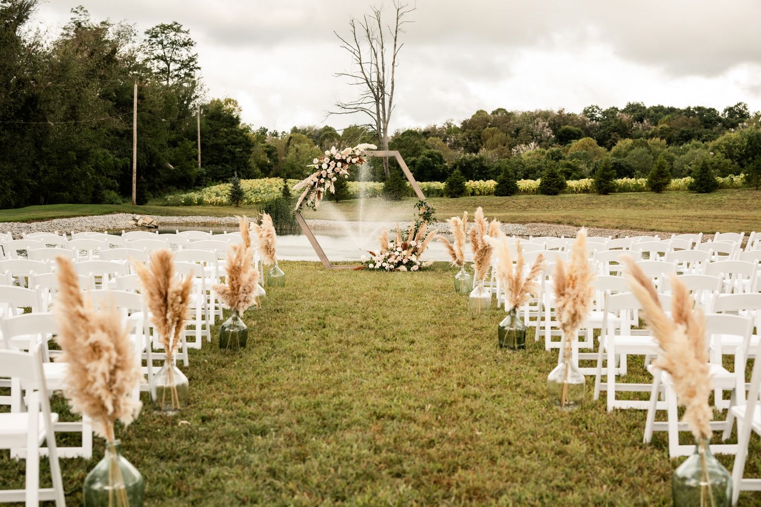 Outdoor wedding ceremony setup with white chairs and pampas grass decorations lining the aisle.