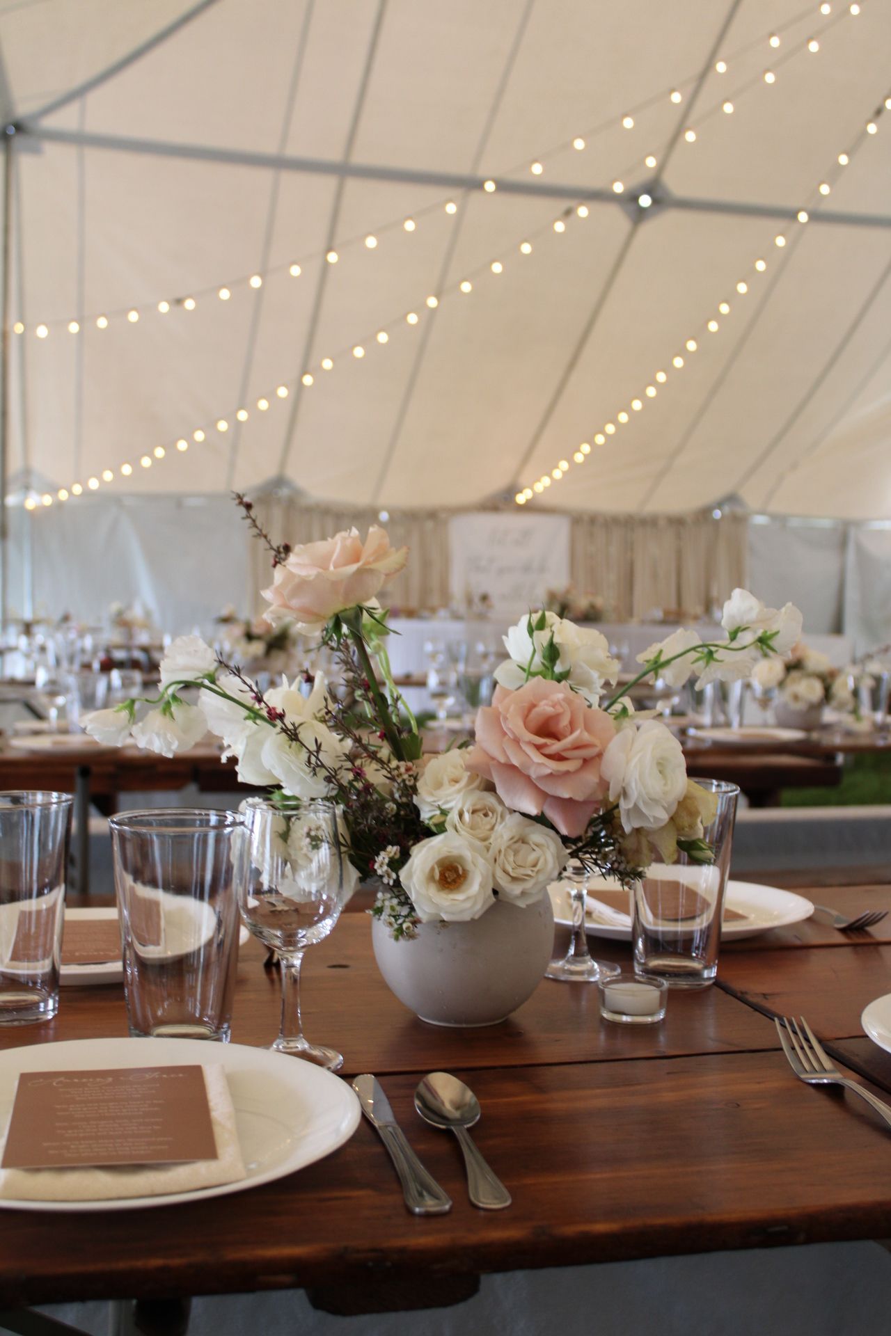 Rustic wedding reception table under tent with string lights and pink rose centerpiece.