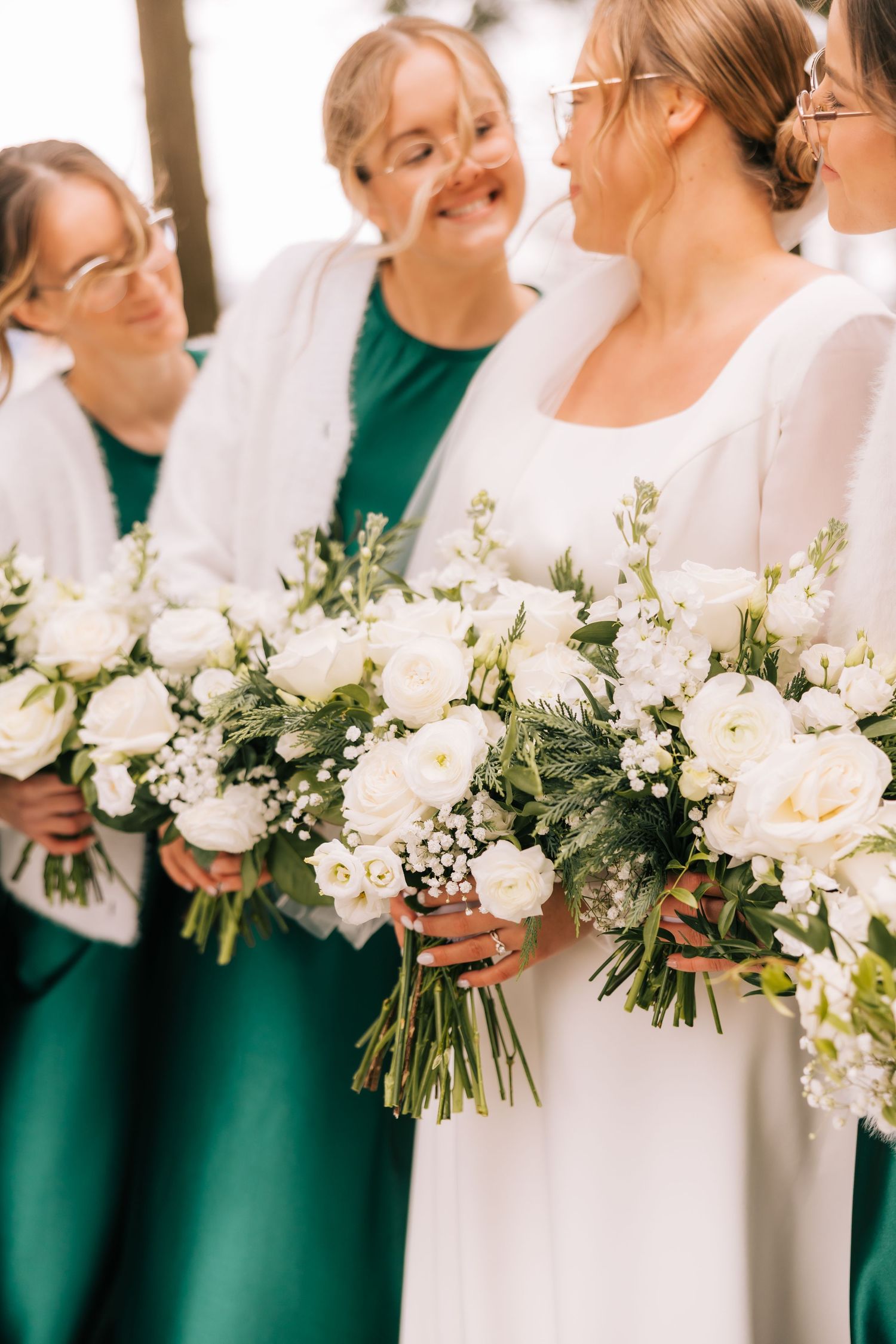 Bridesmaids in emerald green dresses hold elegant white floral bouquets at a wedding ceremony.