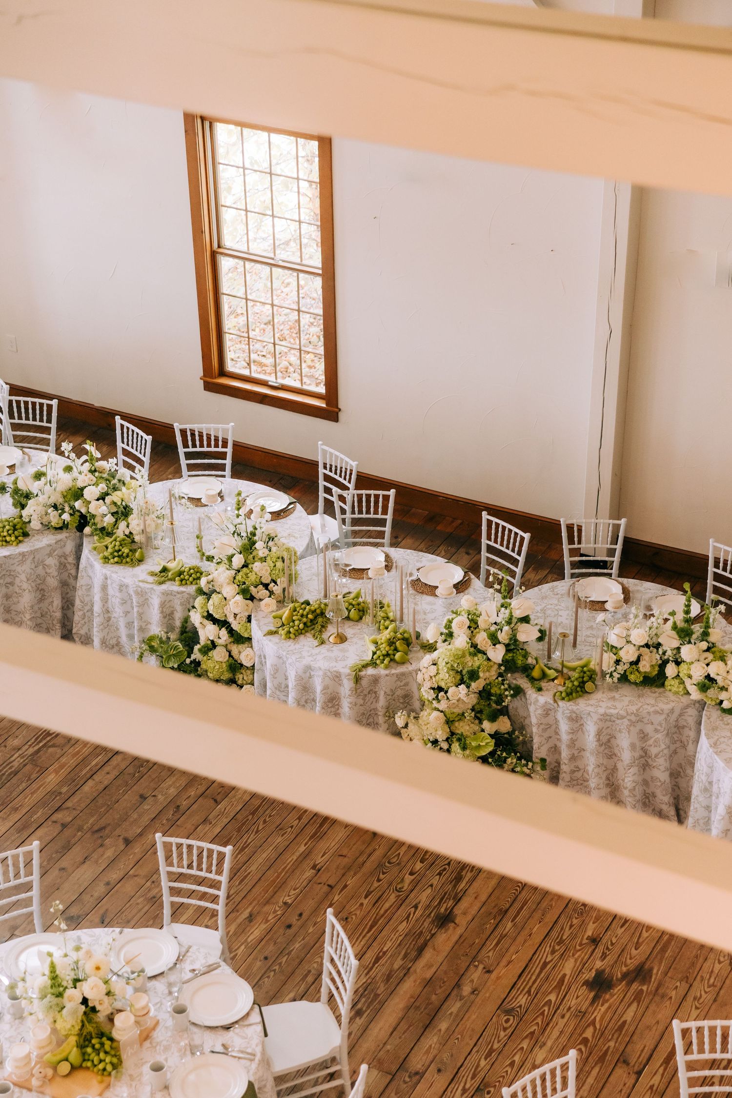 White chiavari chairs and elegant floral centerpieces line rustic wooden tables at an indoor wedding reception venue.