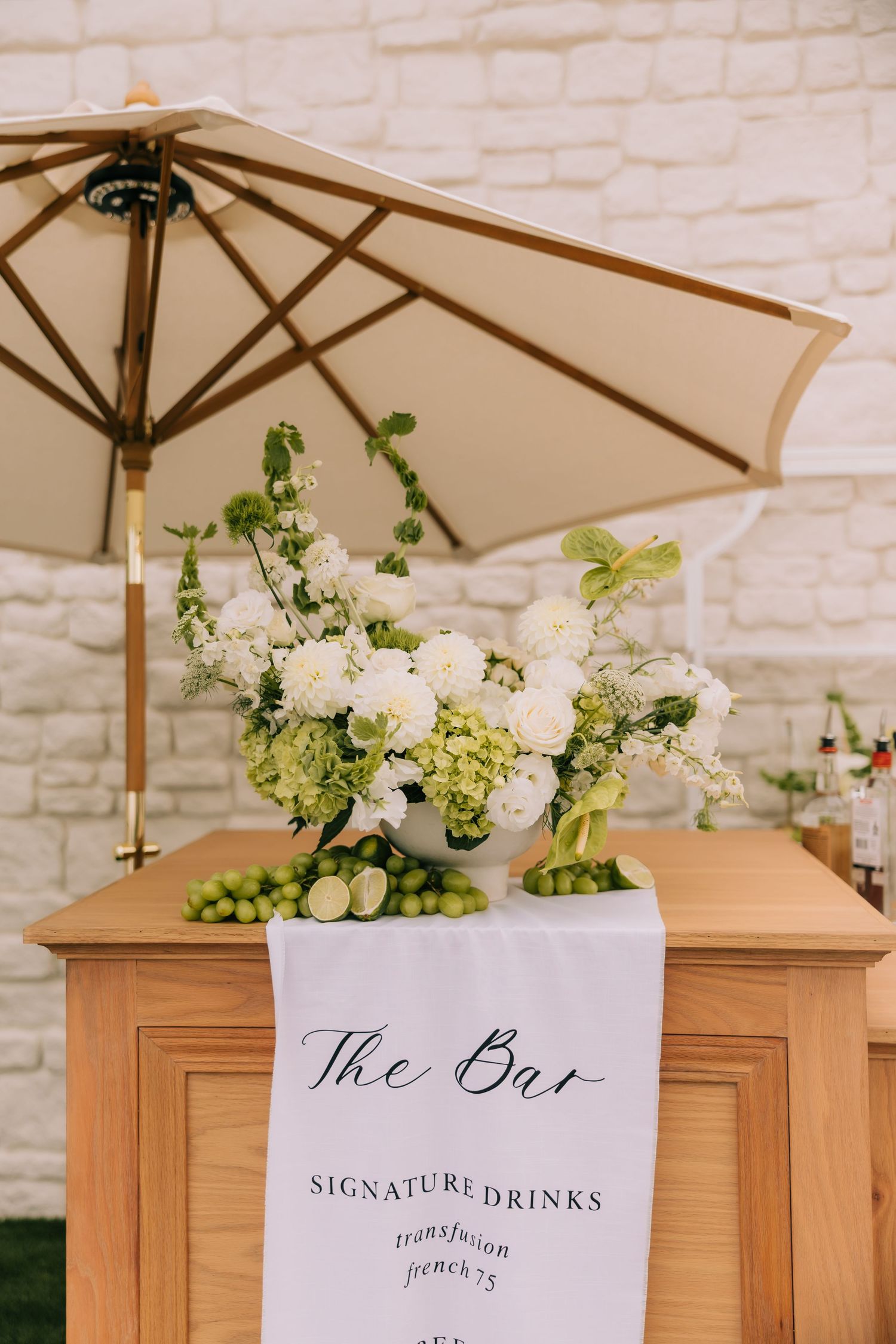 Elegant wedding bar setup with white floral arrangements and signature drinks menu on wooden tables against white brick wall.
