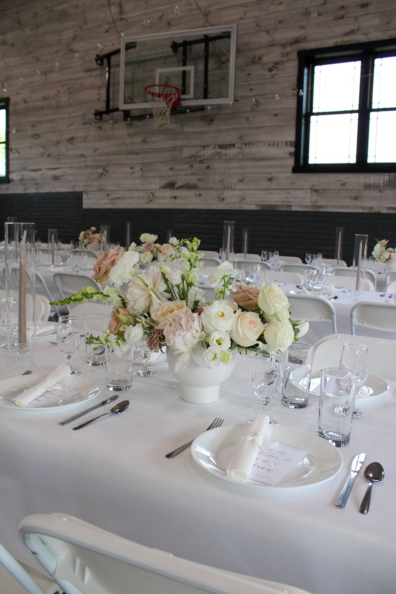 Elegant white wedding reception table settings with floral centerpieces and rustic wood paneled walls in the background.