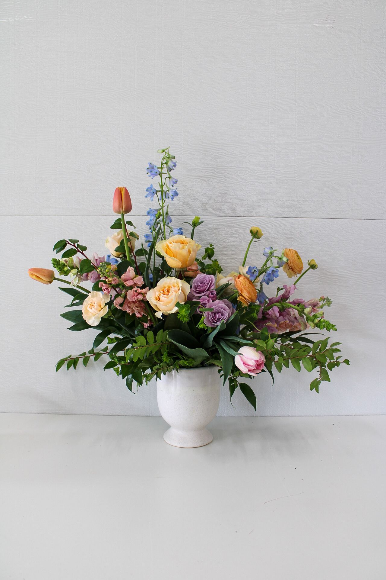 A colorful spring flower arrangement with tulips, roses, and delphinium in a white pedestal vase against a light background.