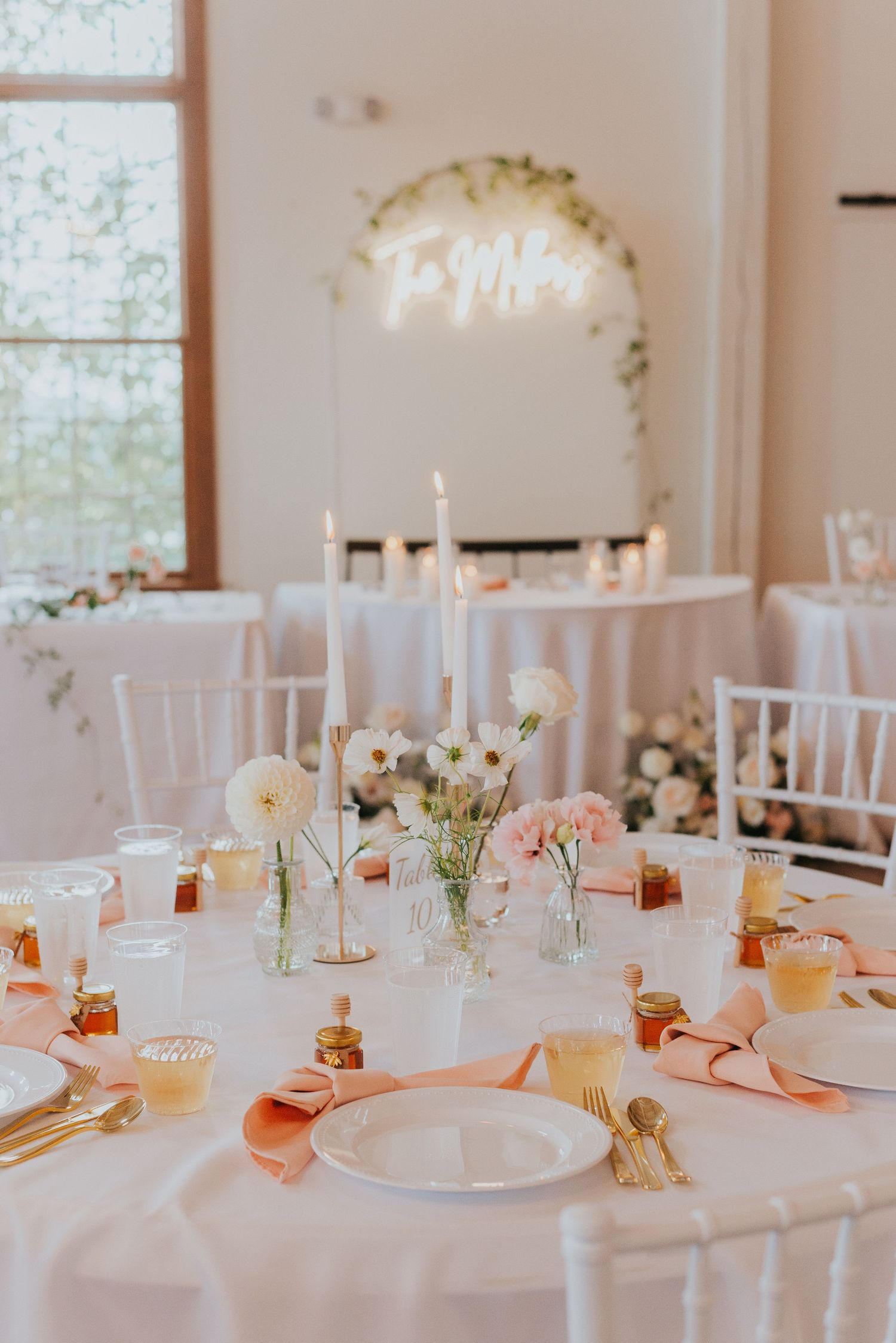 Romantic table setting with white linens, taper candles, and soft floral centerpieces beneath a neon sign.