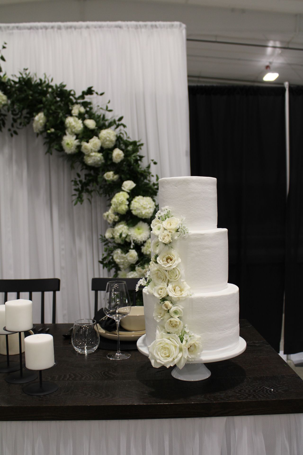 Four-tier white wedding cake decorated with cascading white roses against a white fabric backdrop.