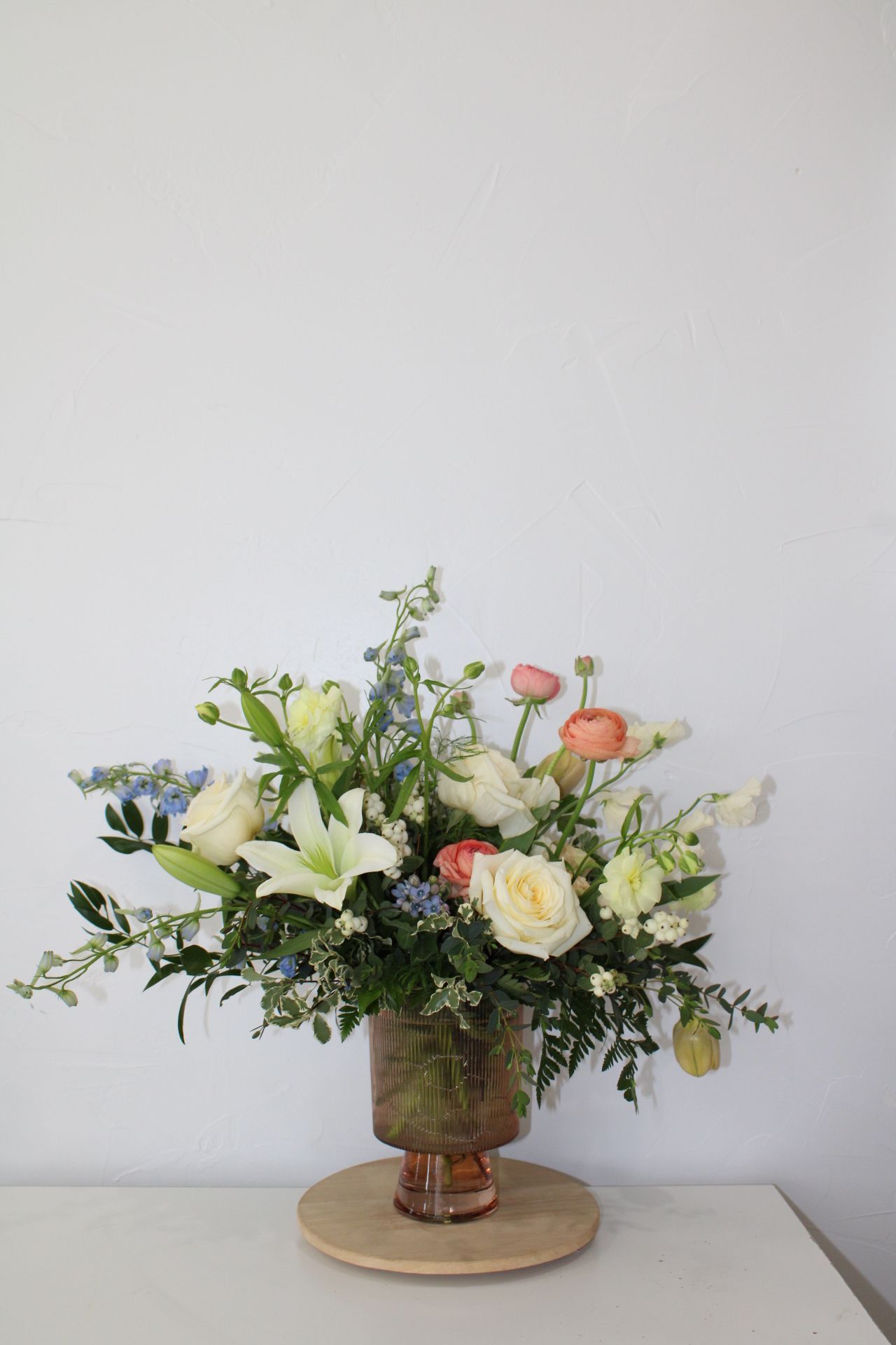 Elegant floral arrangement featuring white lilies and peach roses in a glass vase on a wooden pedestal.