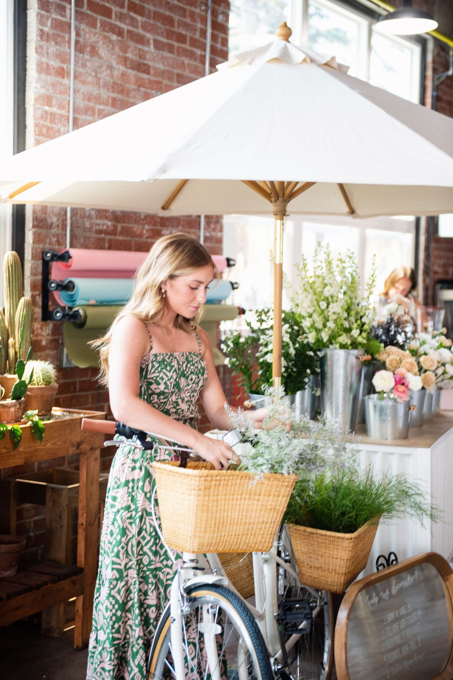 Woman in floral dress arranging flowers near vintage bicycle under white market umbrella.