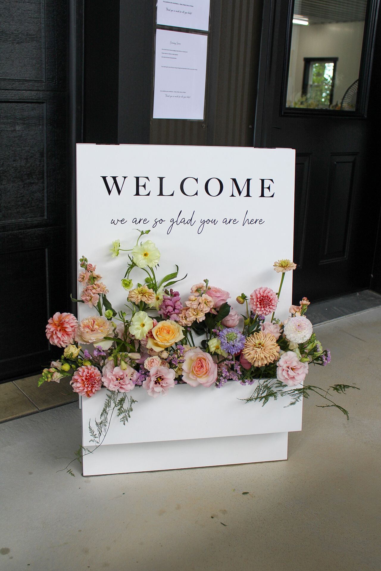 A white welcome sign decorated with pink, peach and yellow floral arrangements including roses and dahlias.