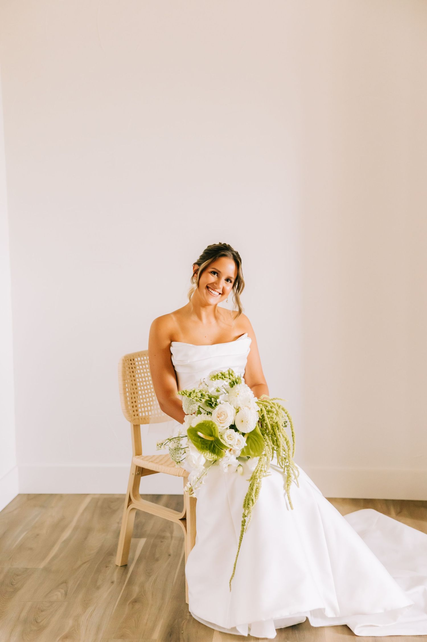 Bride in elegant white wedding gown poses gracefully on wooden chair against neutral backdrop in minimalist setting.