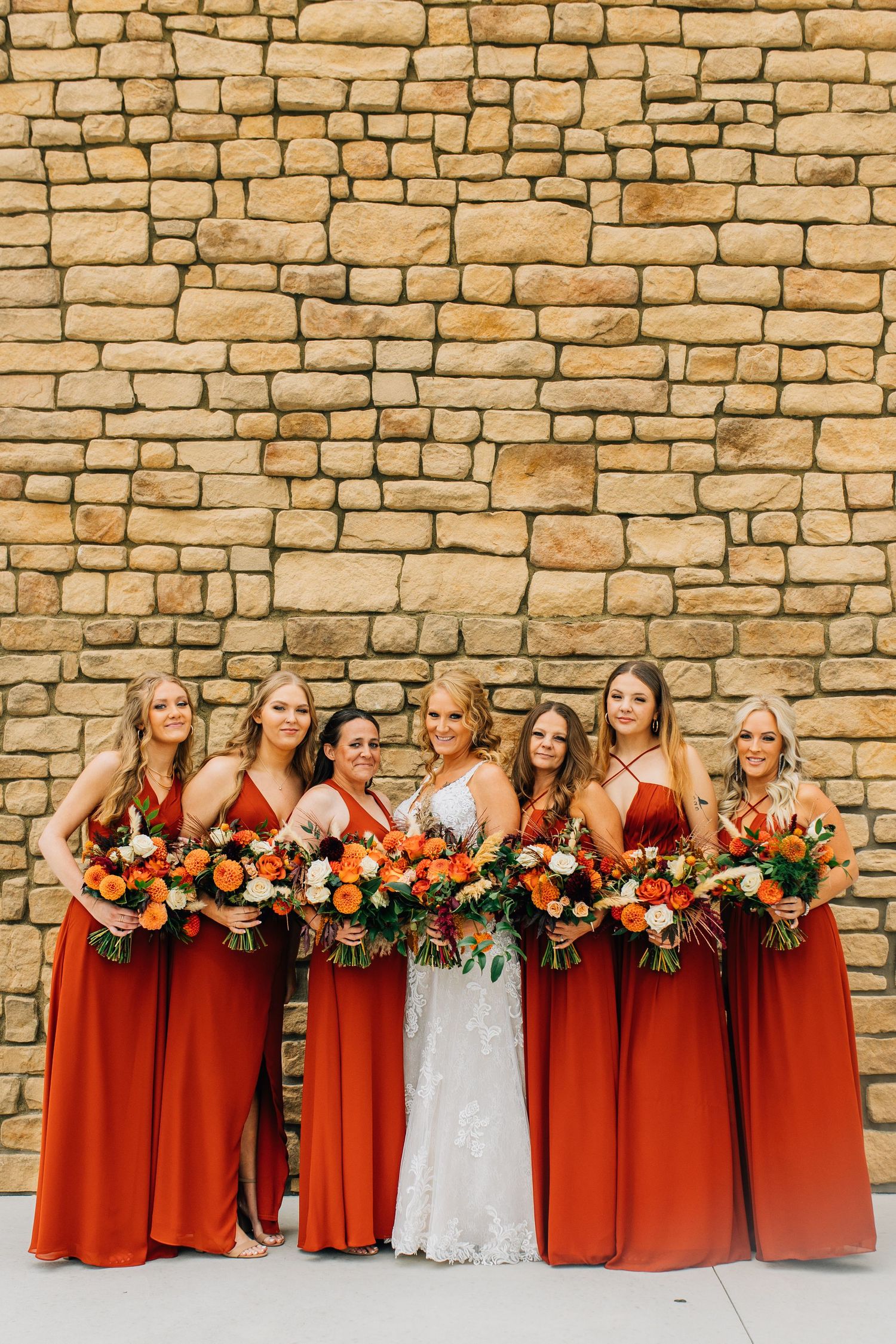 Bridal party in matching rust-colored gowns pose with bouquets against a stone wall backdrop.