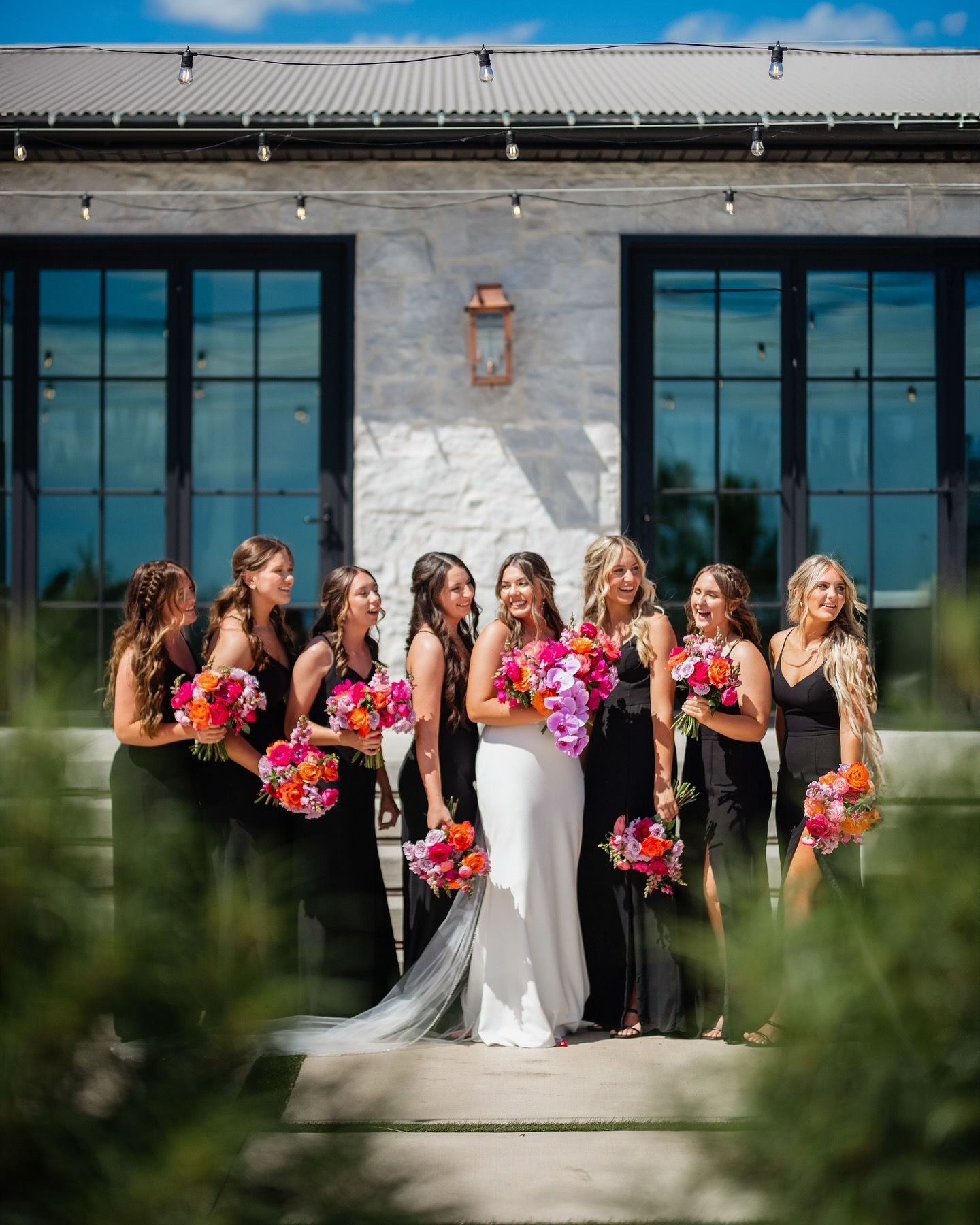 Bridal party in black dresses with colorful bouquets posed against modern building.