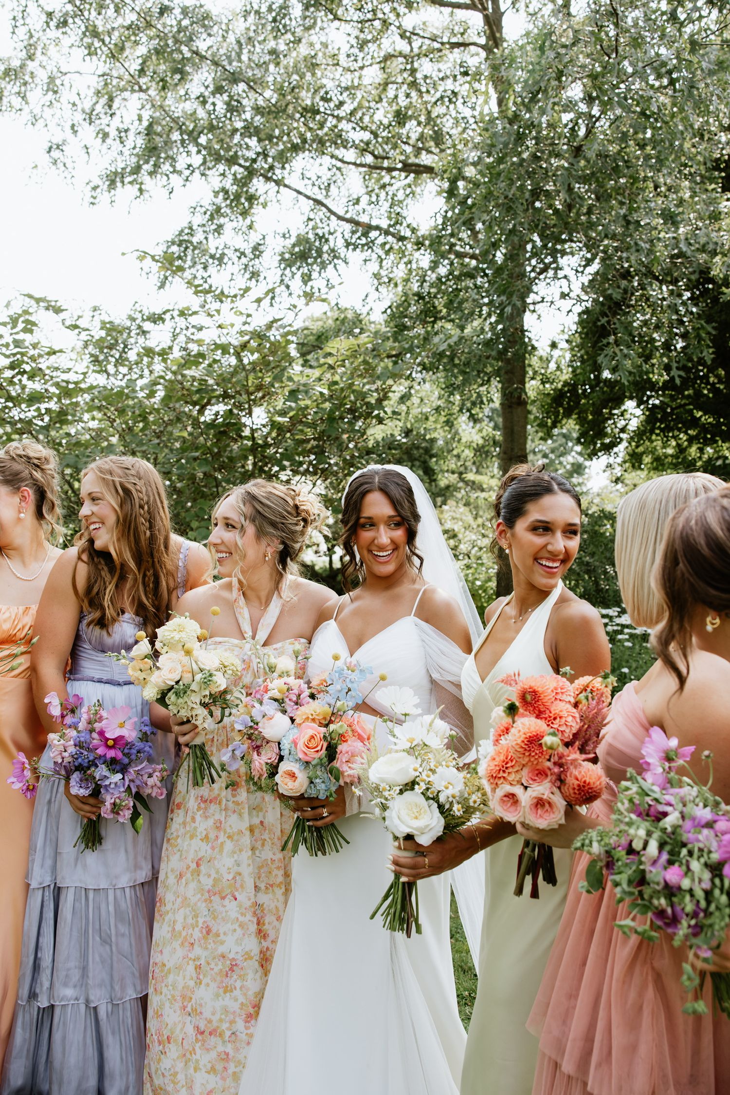 Wedding party featuring bridesmaids in mismatched pastel dresses holding colorful floral bouquets in an outdoor garden setting.