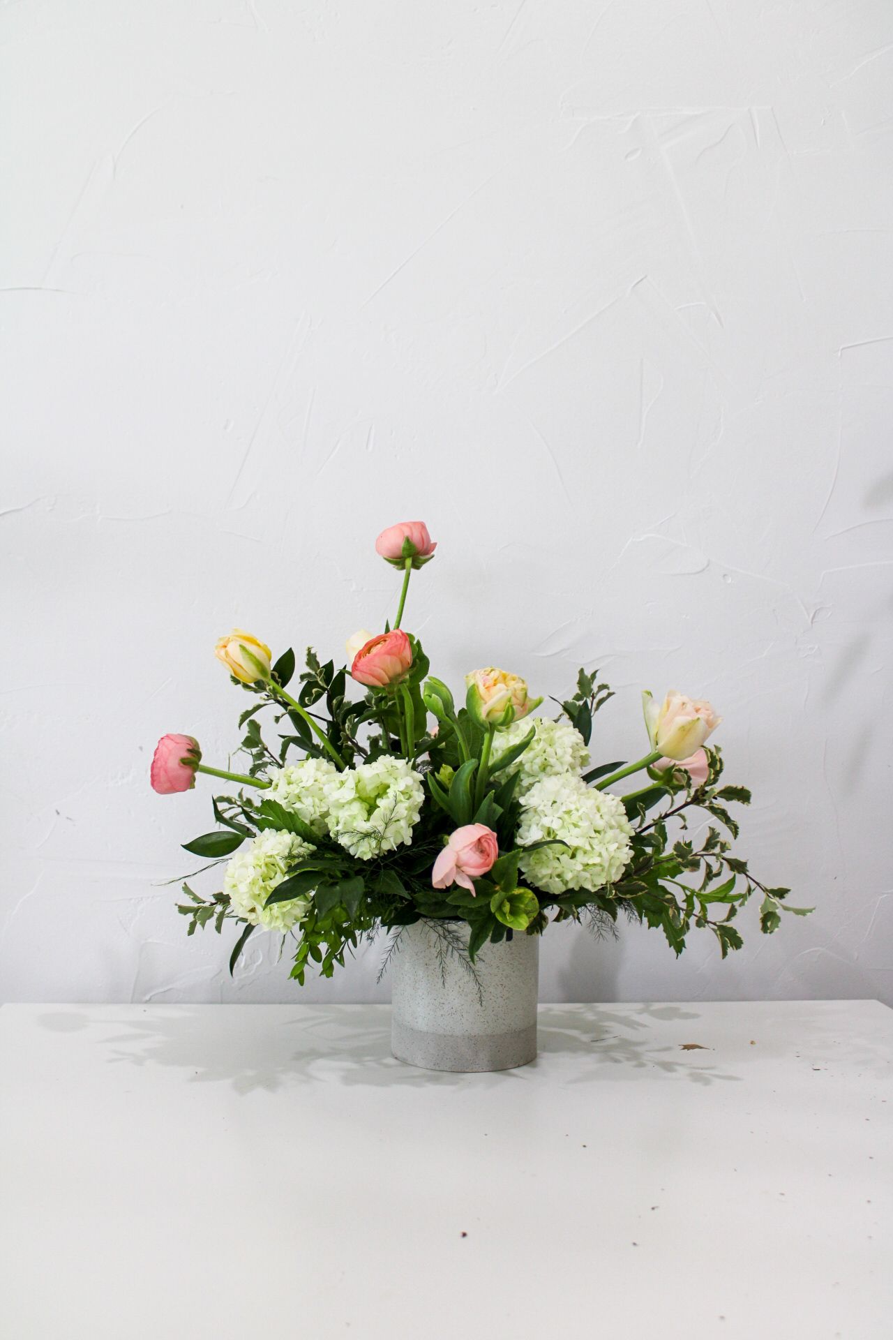Spring arrangement featuring pink ranunculus, white hydrangeas and yellow blooms in gray container.