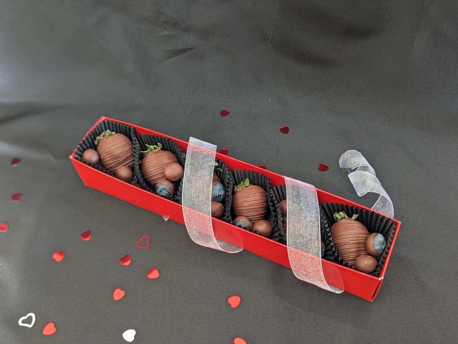 Chocolate truffles arranged in a black box with red ribbon and scattered heart decorations on gray surface.