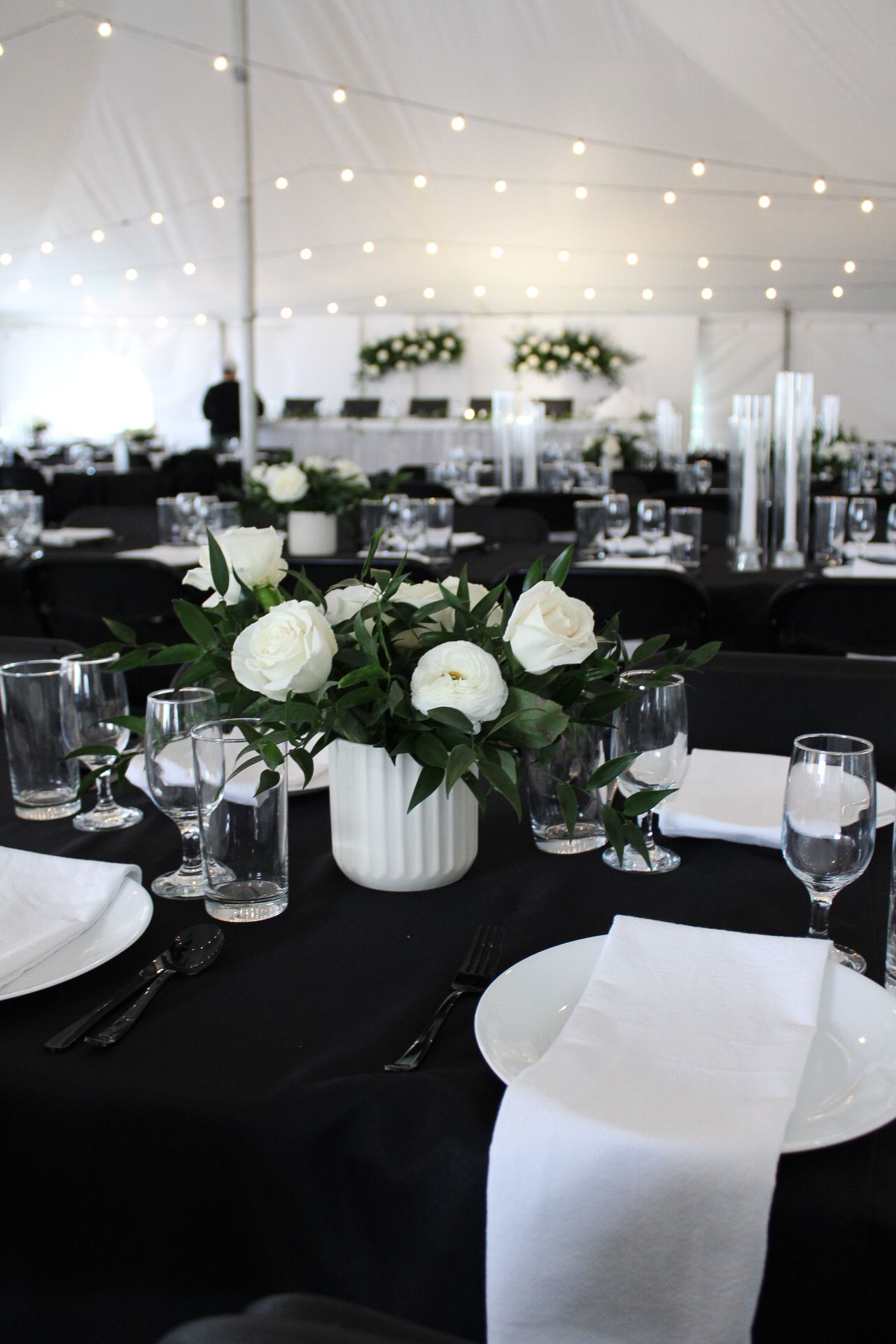 Elegant wedding reception tables with black linens, white place settings, and string lights overhead in a tent venue.