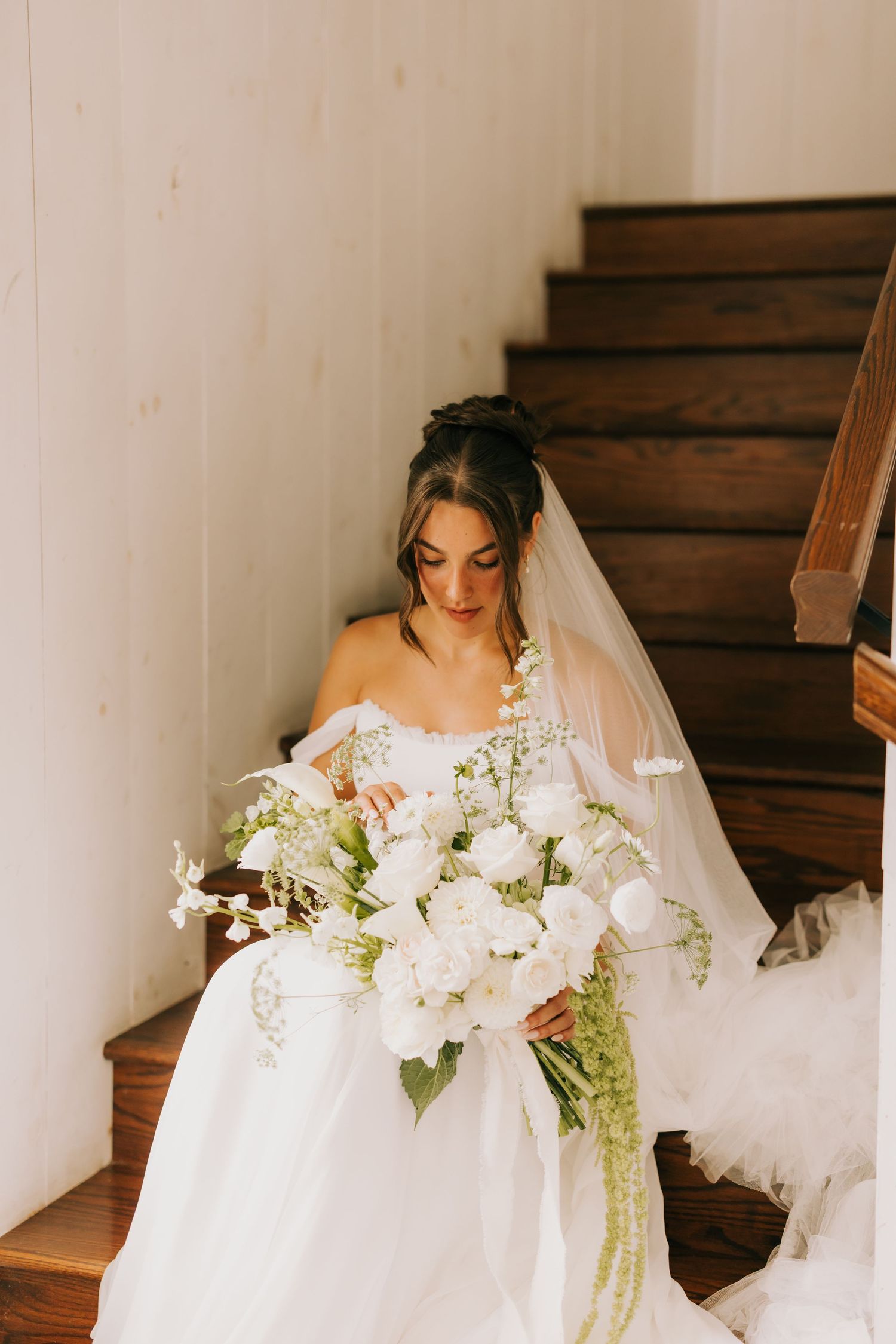 A bride in a flowing white wedding dress sits gracefully on wooden stairs surrounded by delicate floral arrangements.