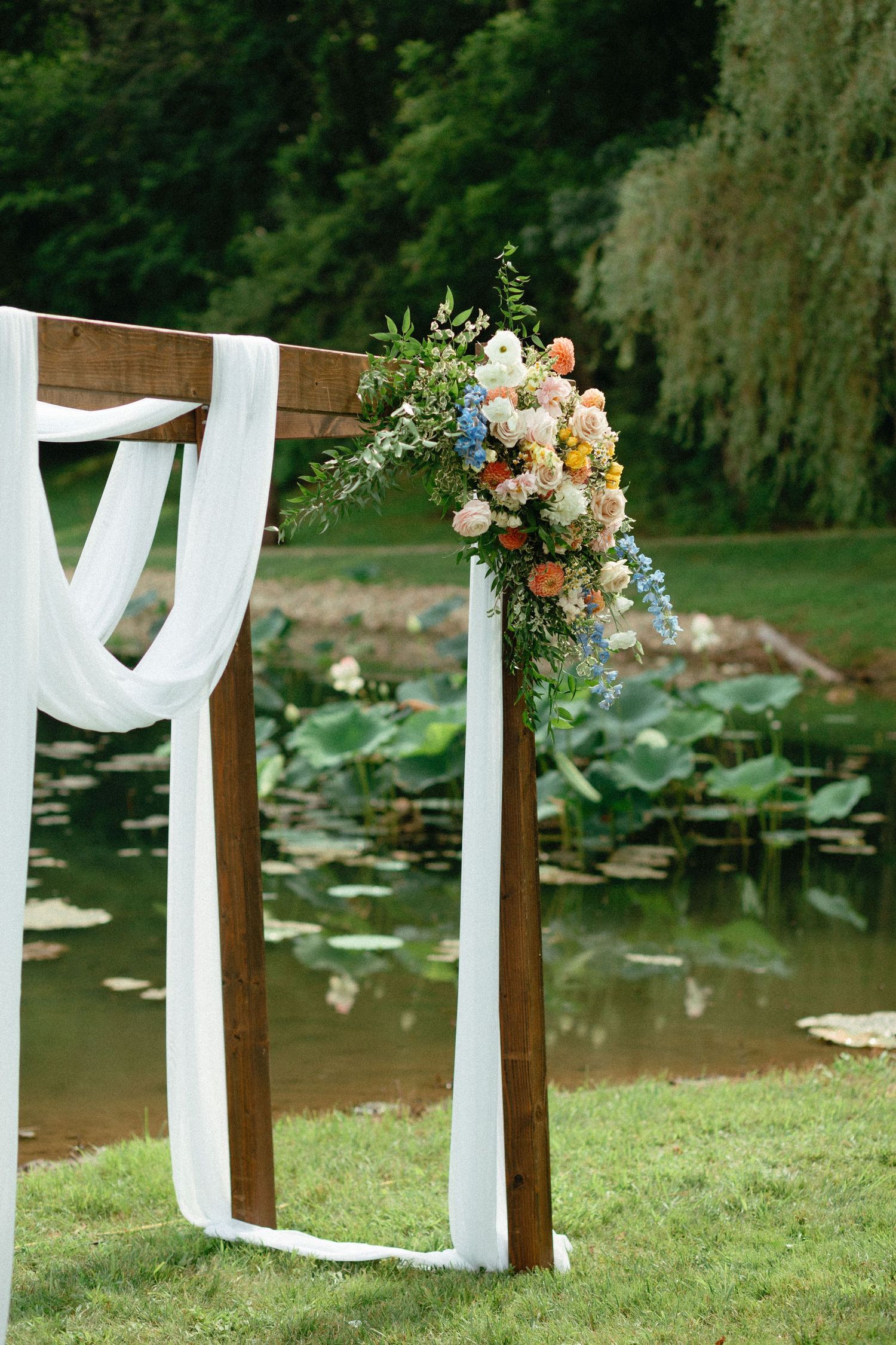 White fabric and floral decorations adorn wooden posts along a pond with lily pads for an outdoor wedding ceremony.