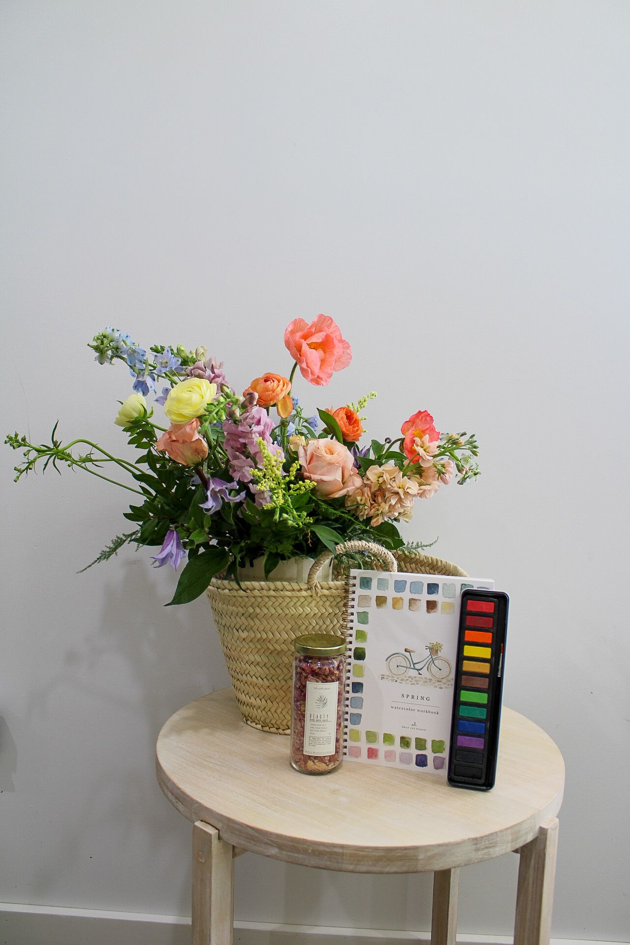 Colorful floral arrangement in a wicker basket on a wooden side table with watercolor paints and a small jar.