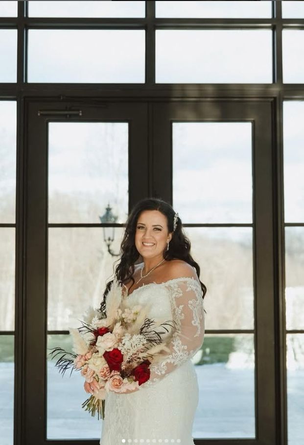 Winter wedding portrait in front of large windows with bridal bouquet featuring red roses.