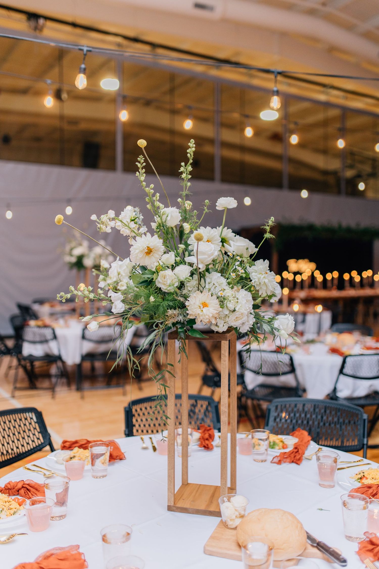 Tall white floral centerpiece on modern wooden stand with coral accents on round reception table.