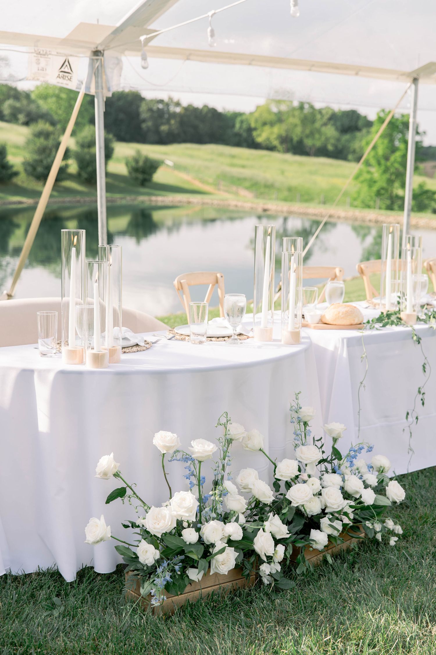 Outdoor wedding reception setup with white linens and floral arrangements overlooking a serene pond landscape.
