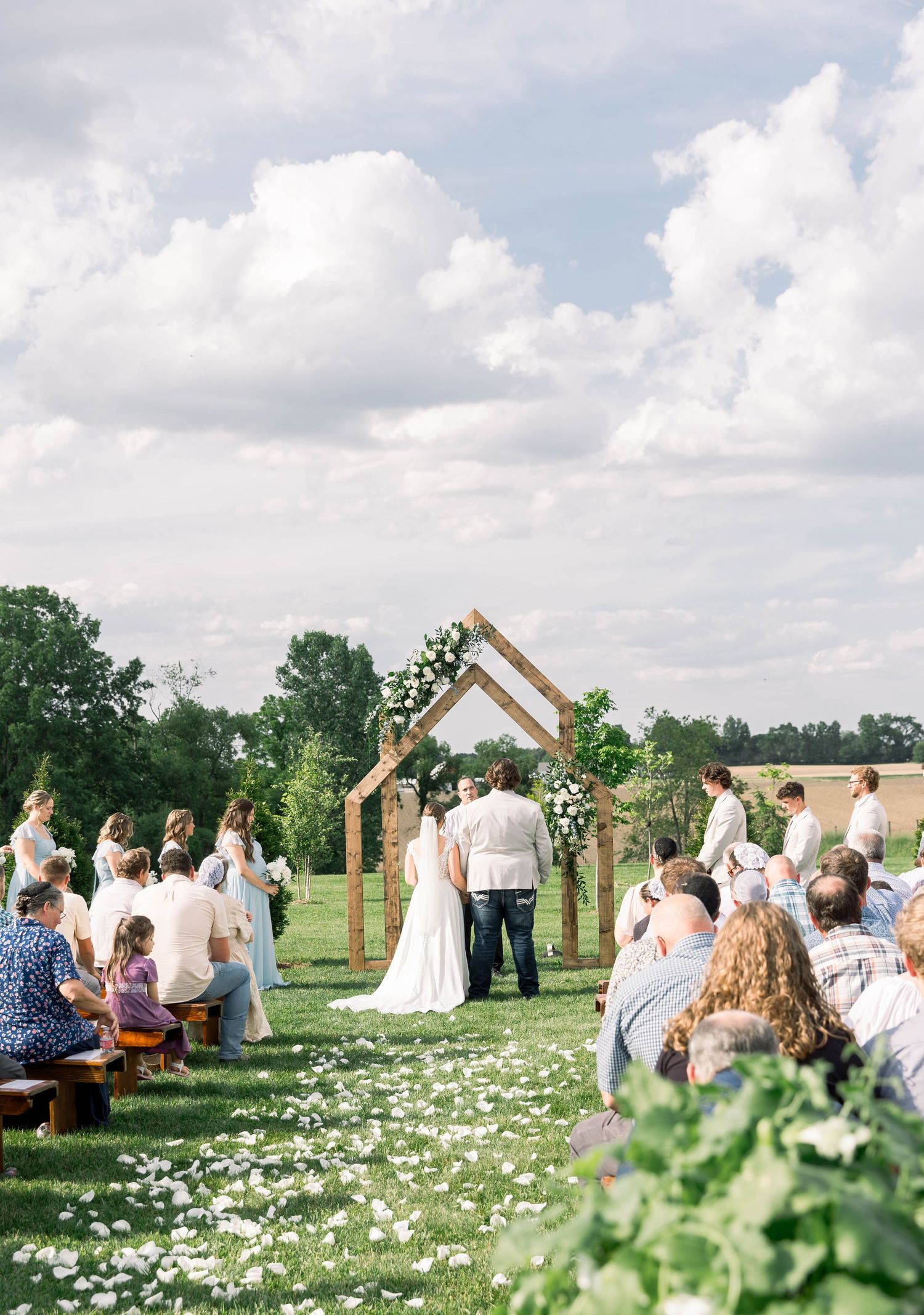 Outdoor wedding ceremony under wooden arch with flower petals scattered along aisle and wooden bench seating.