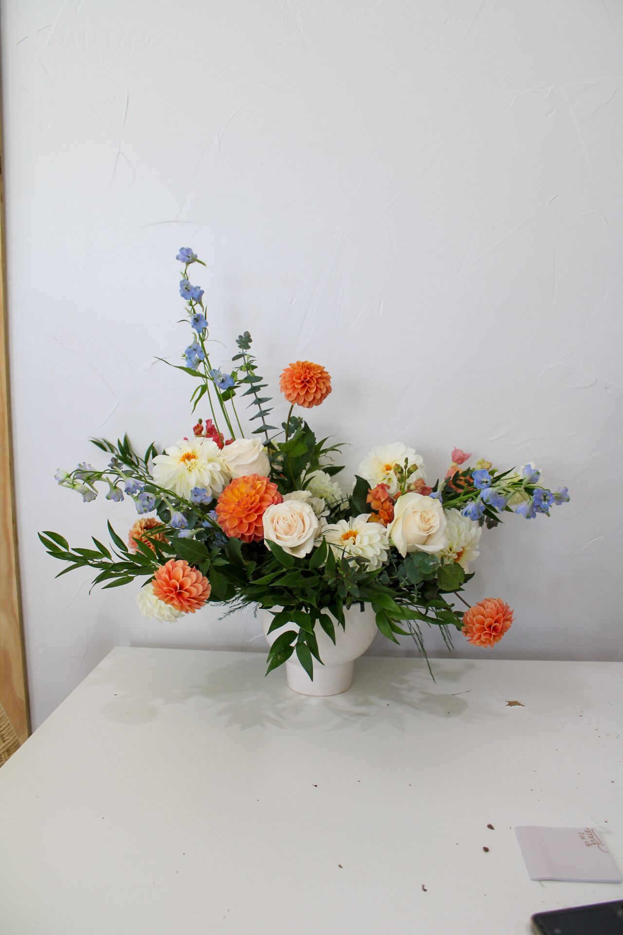 Fresh white roses and orange carnations with blue delphinium in a white ceramic vase.