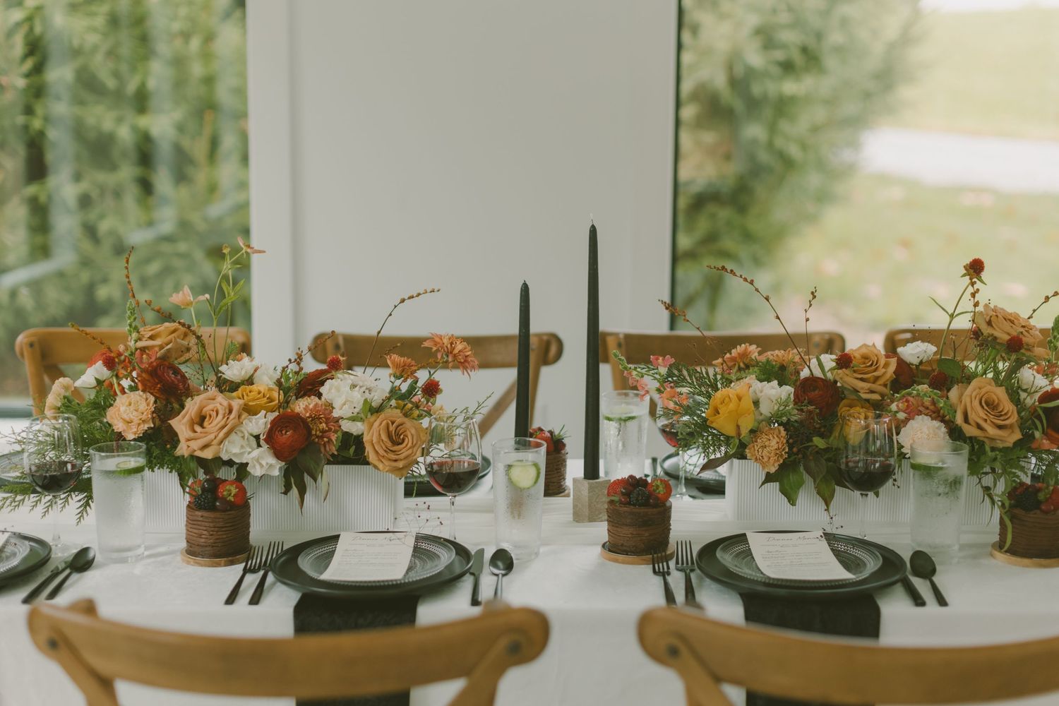 Wedding table setting with black plates, tall candles, and rustic floral arrangements in wooden vessels.