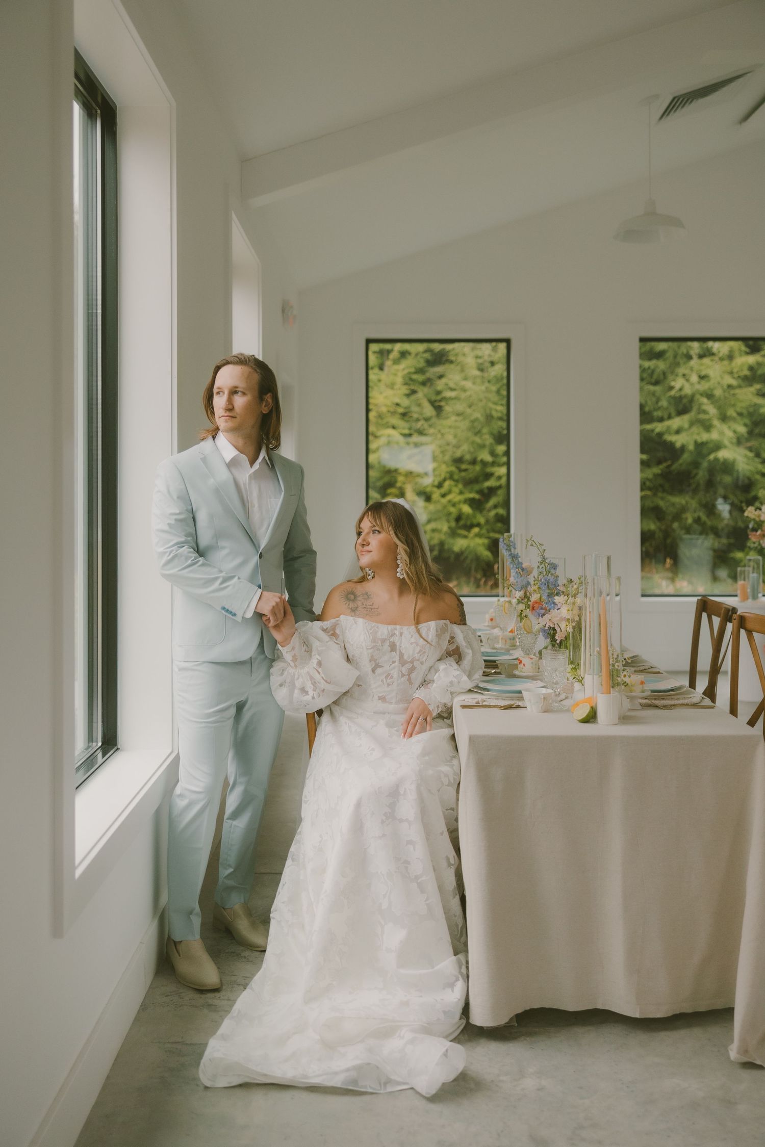 A couple in elegant wedding attire poses by a window in a bright white room with a formal table setting.