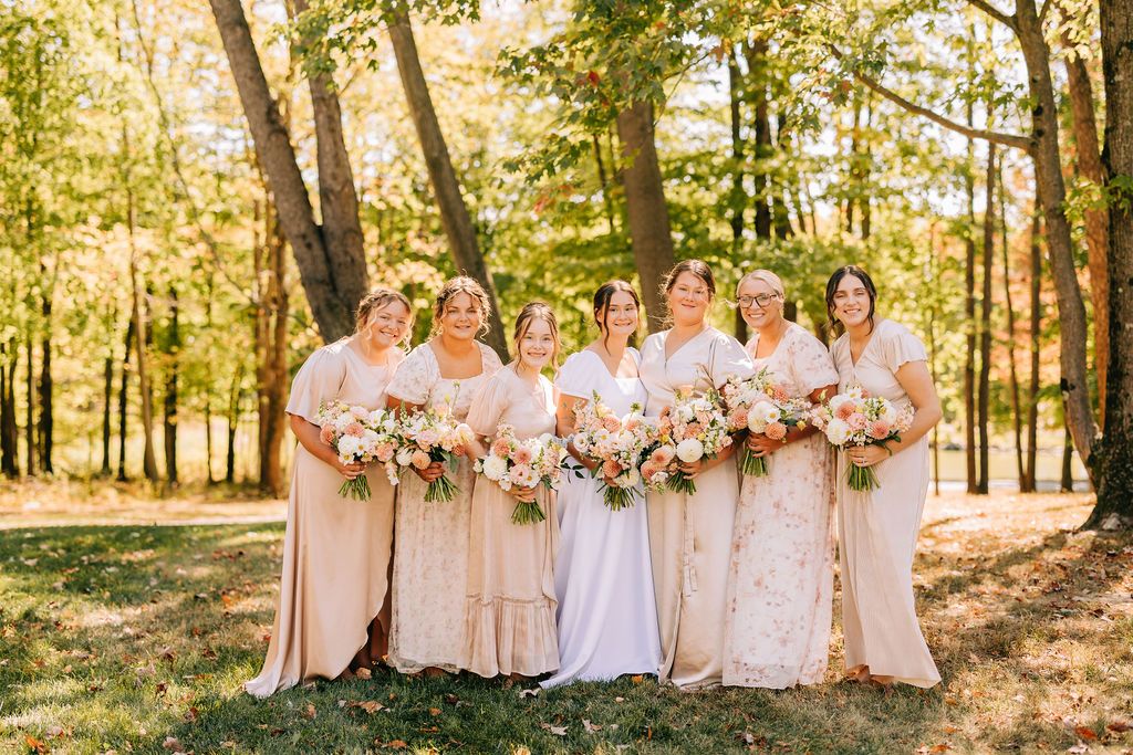 A wedding party in blush pink dresses stands together in a sunlit wooded area during golden hour.