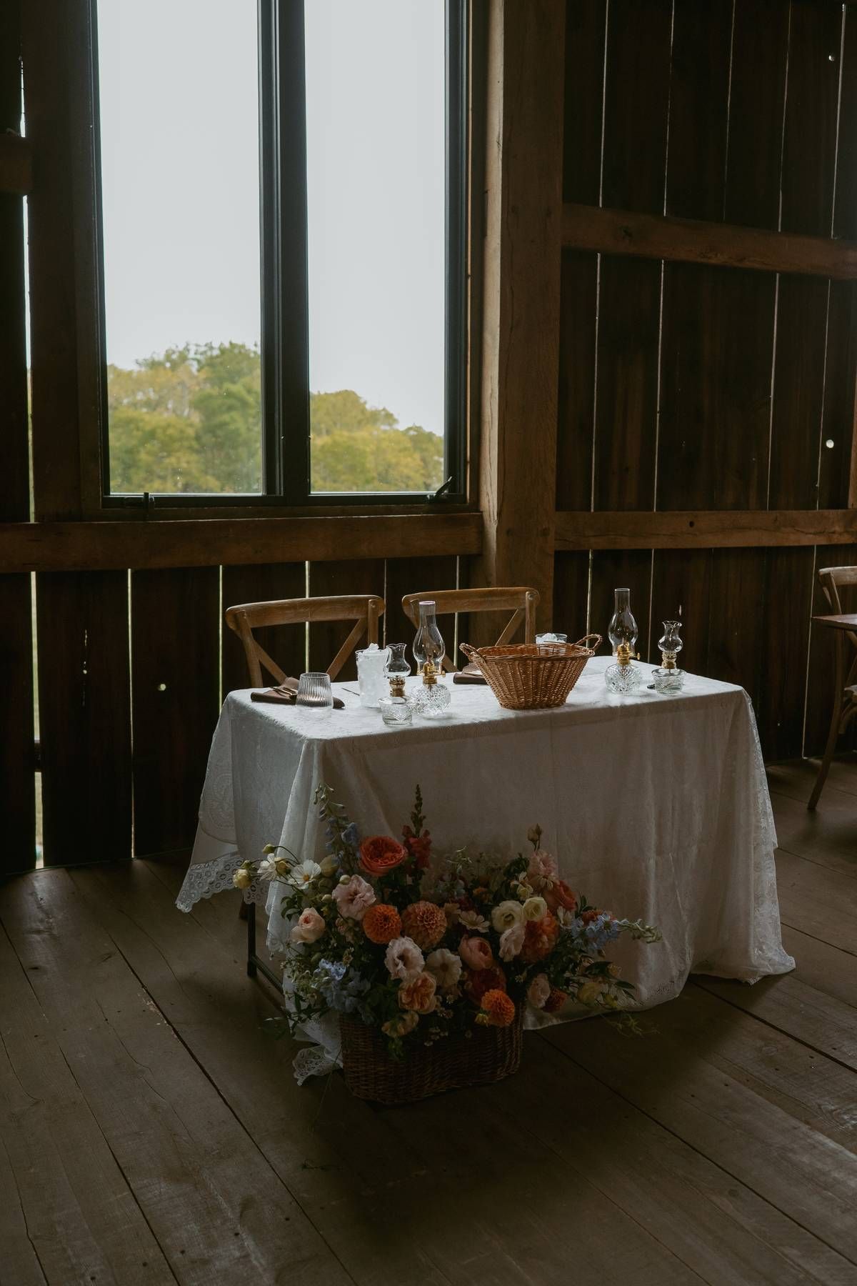 Rustic barn reception tables with white linens and floral centerpieces set against large windows with natural lighting.
