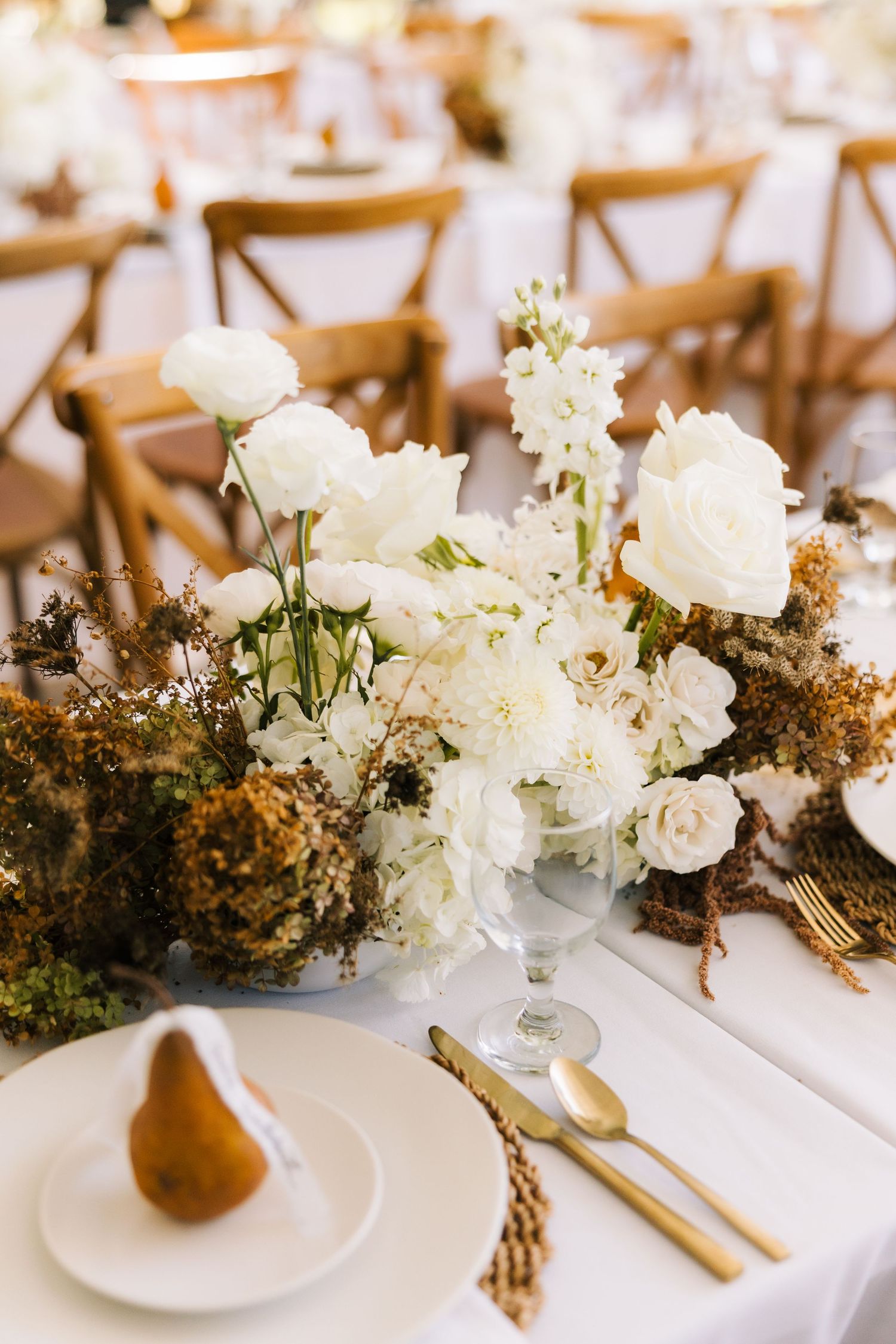 White floral centerpieces with dried foliage create an elegant tablescape at a wedding reception with gold cutlery.