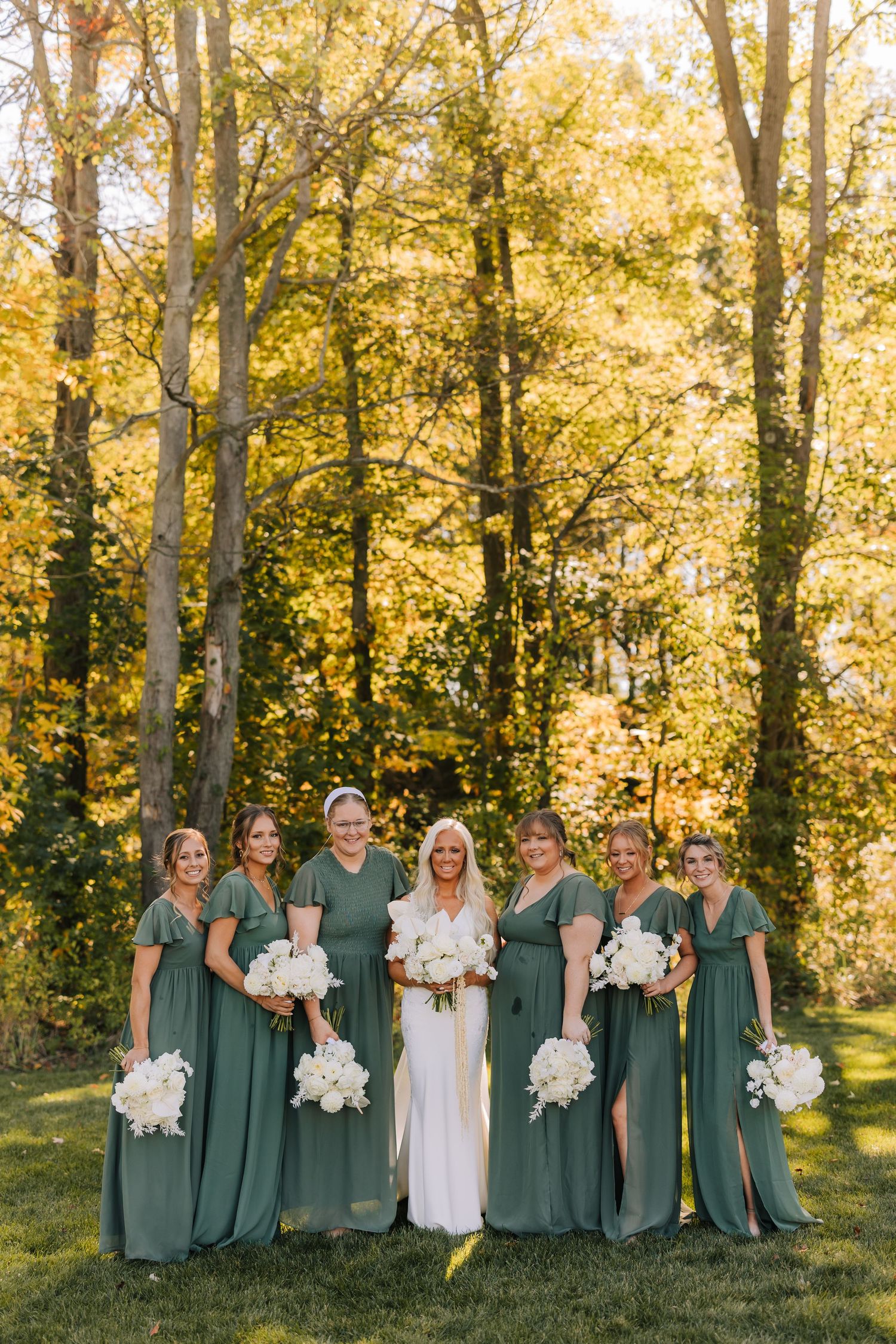 Wedding party wearing sage green dresses and white floral bouquets poses together in front of autumn trees at sunset.
