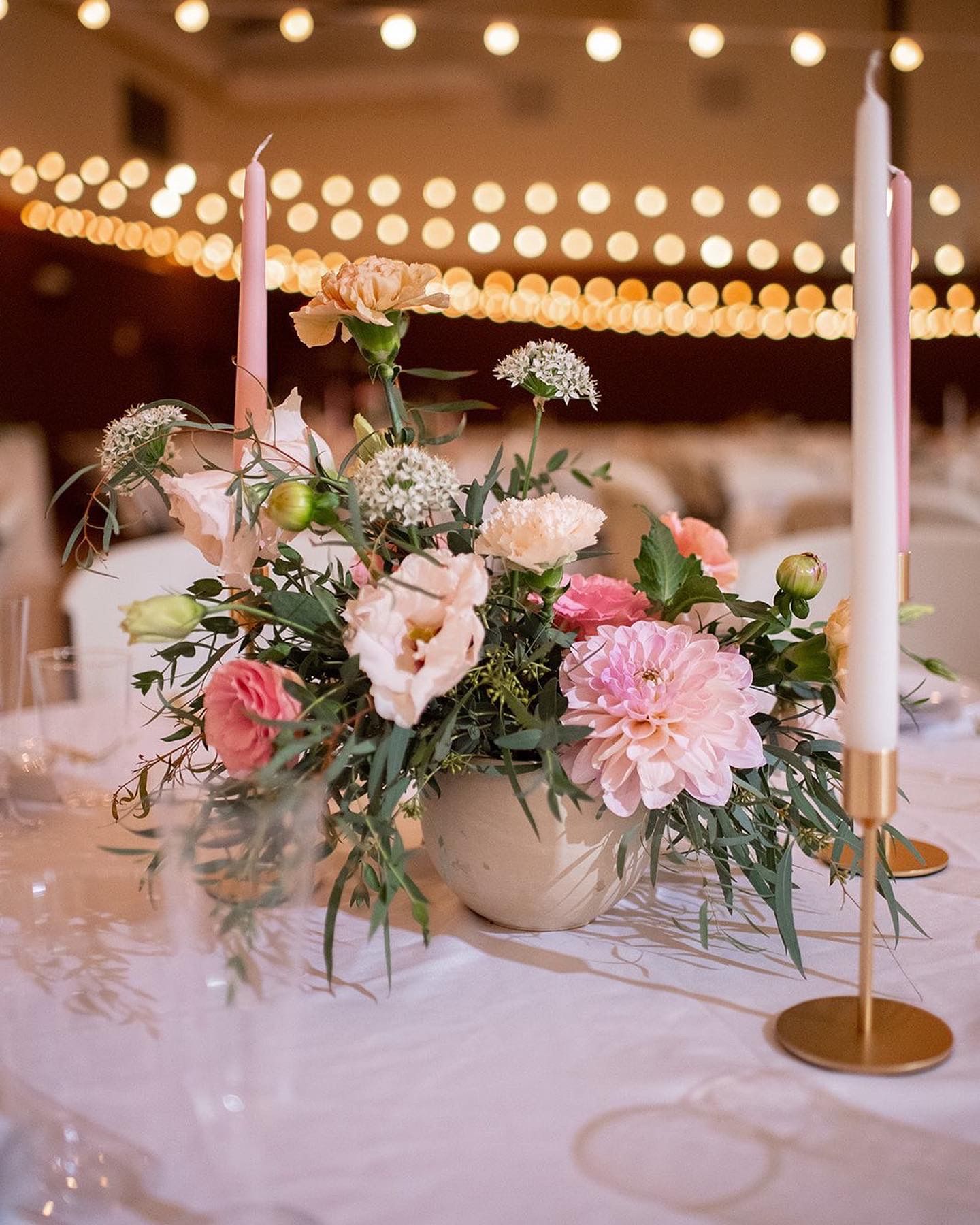 Romantic centerpiece with pink flowers, greenery and tapered candles on white tablecloth.